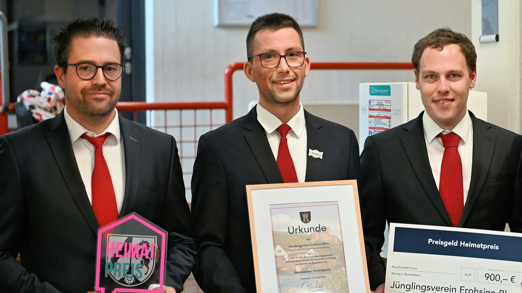 Drei Männer in dunklen Jacketts und roten Krawatten halten den Heimatpreis, die Urkunde und den Scheck in ihren Händen.
