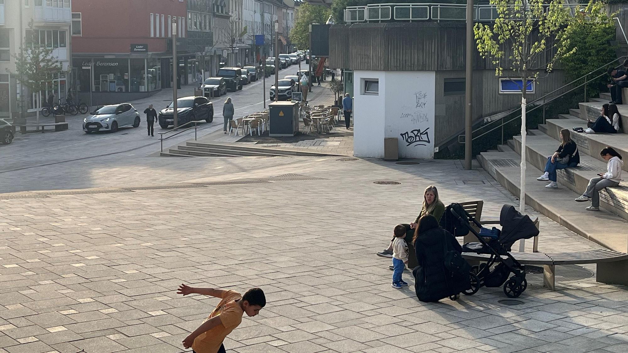 Menschen sitzen auf einer Bank, ein Junge spielt Fußball.