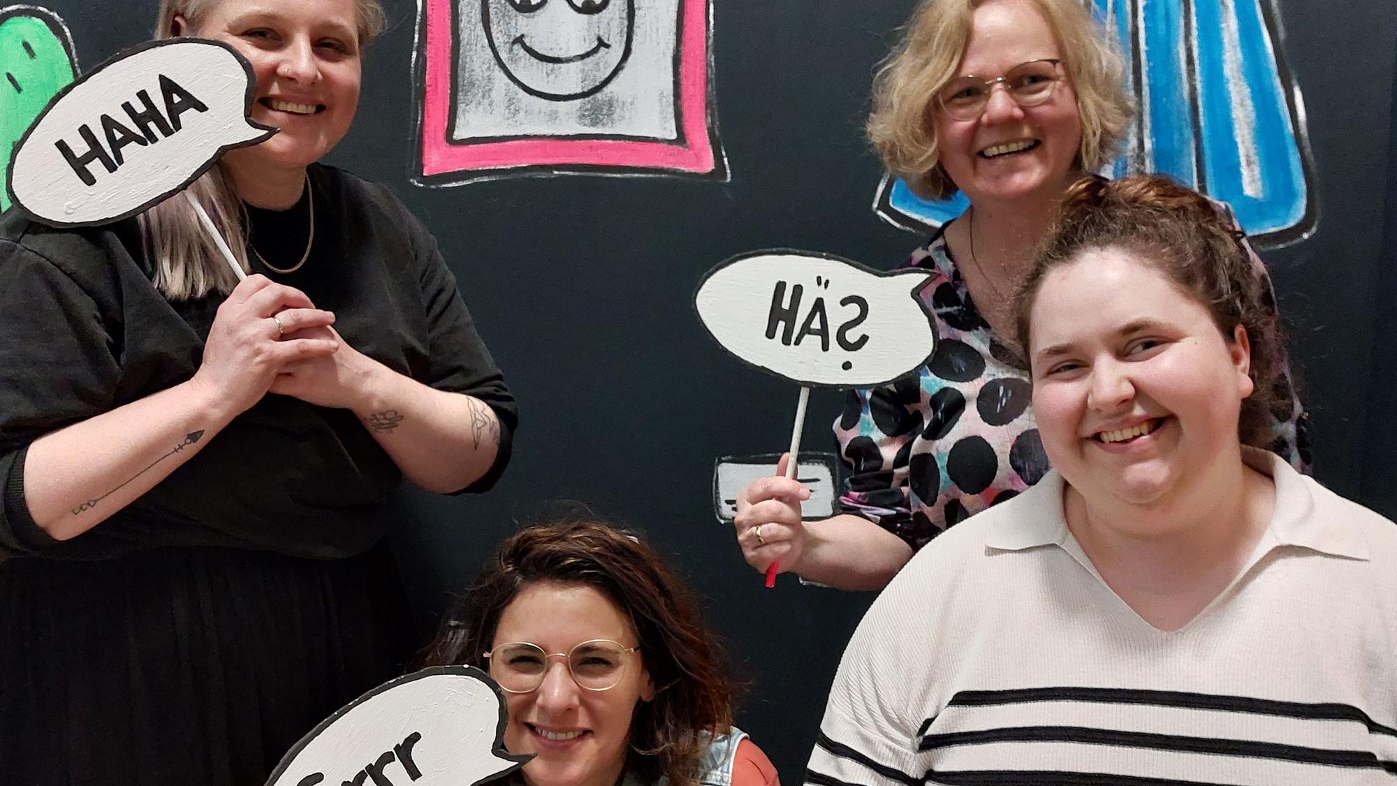 Vier Frauen halten Comicsprechblasen an Holzstäben.