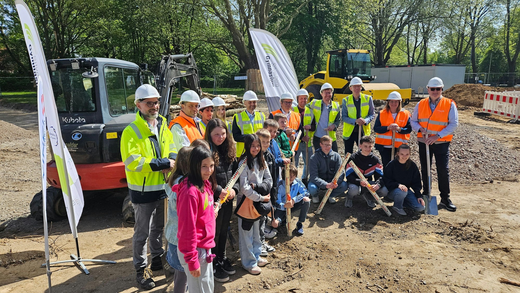 Zu sehen sind Menschen mit Helmen und Warnwesten sowie Schülerinnen und Schüler auf einer Baustelle.