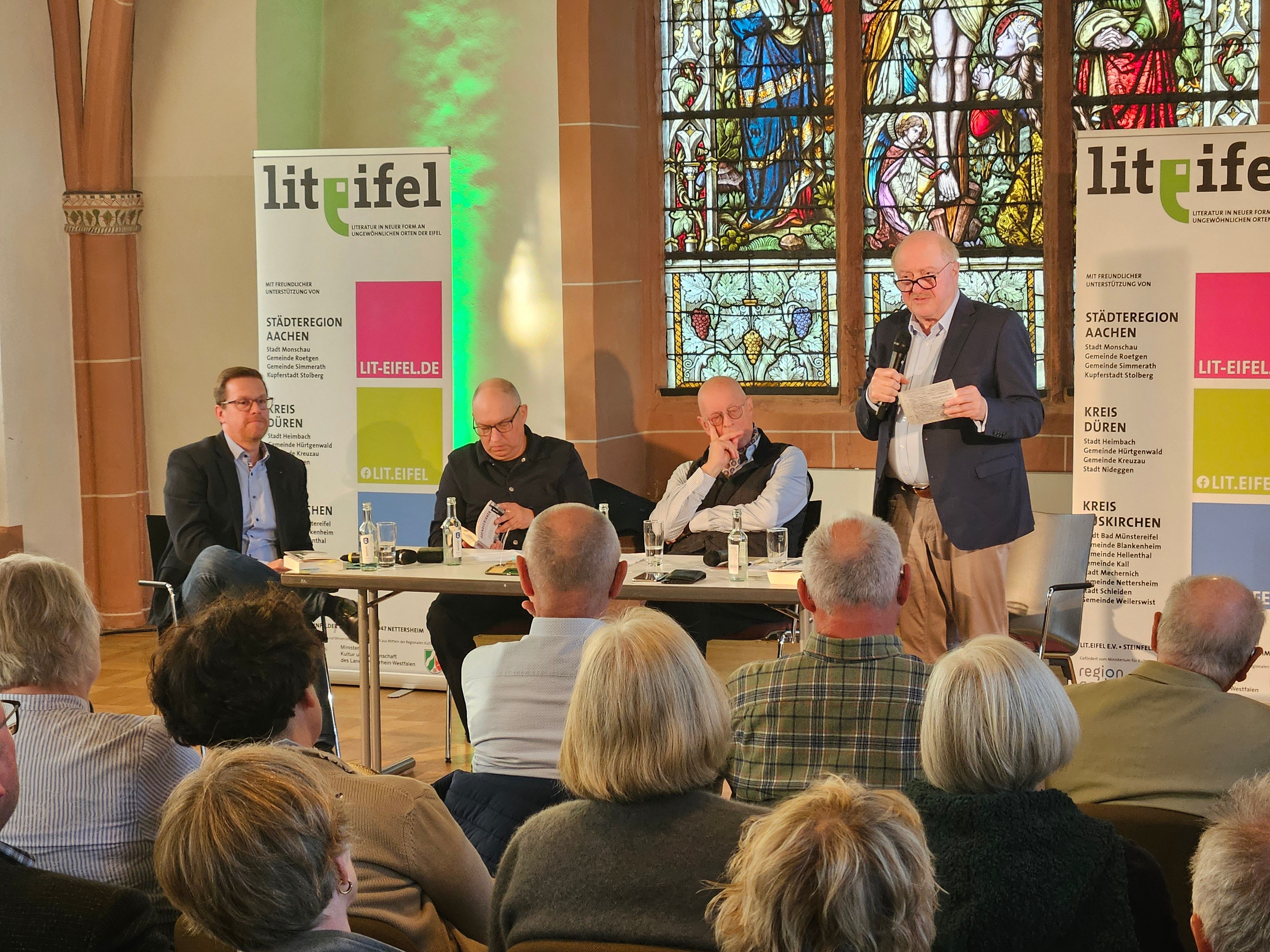 Ein Mann steht rechts mit einem Mikro, daneben sitzen drei Männer an einem Tisch.