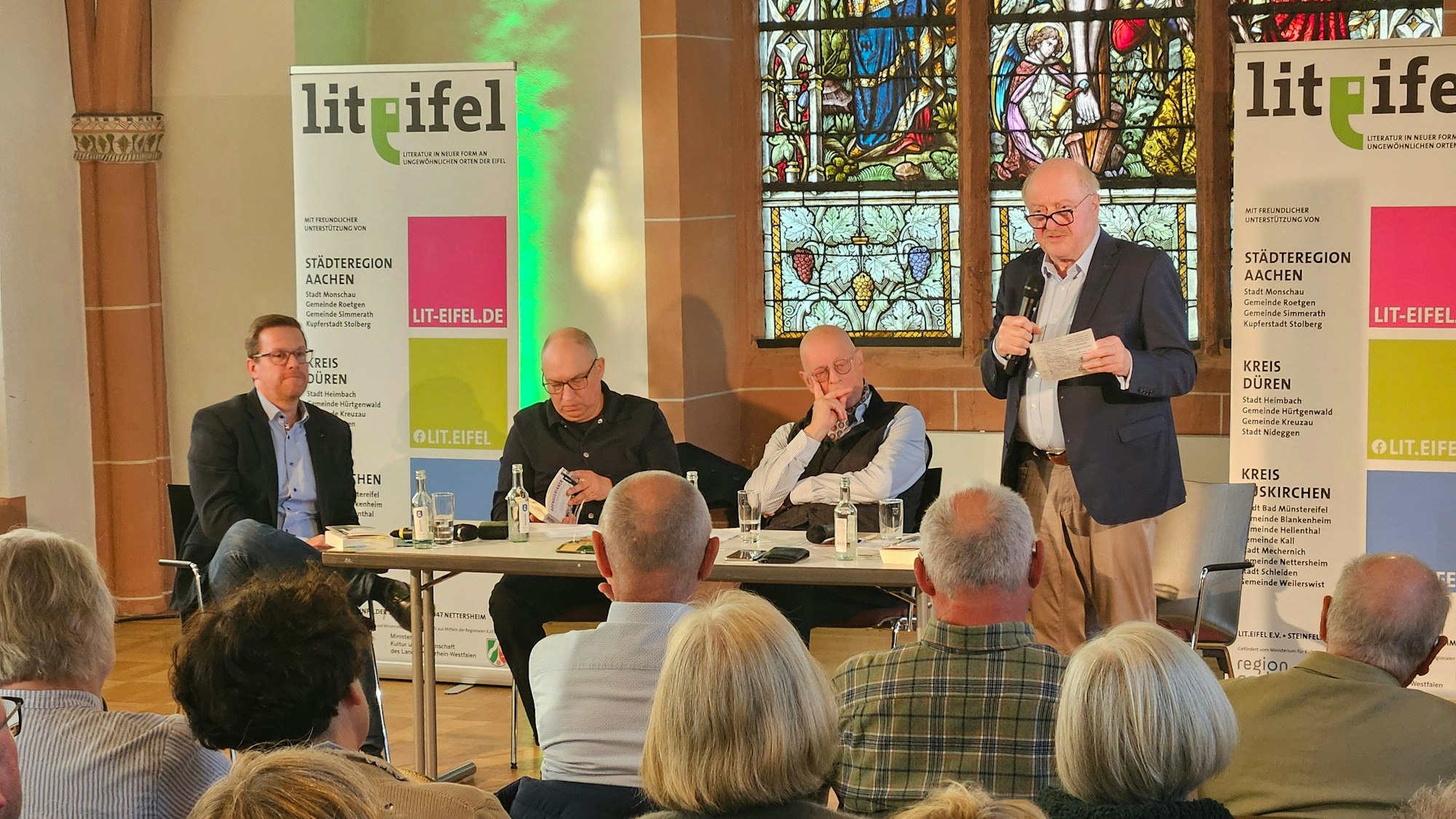 Ein Mann steht rechts mit einem Mikro, daneben sitzen drei Männer an einem Tisch.