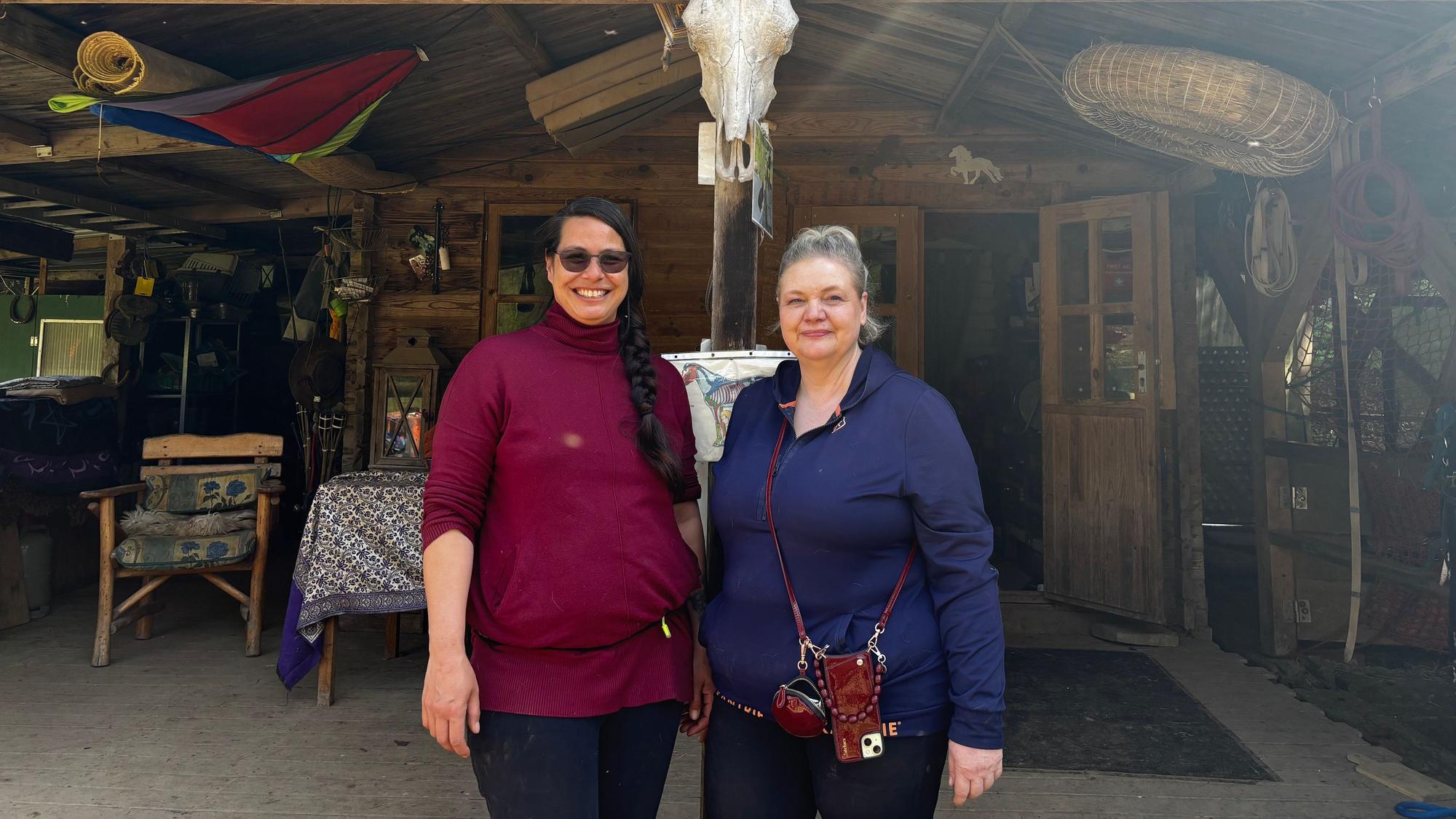 Zwei Frauen stehen nebeneinander und blicken lächelnd in die Kamera. Im Hintergrund ist die Ranch zu sehen.