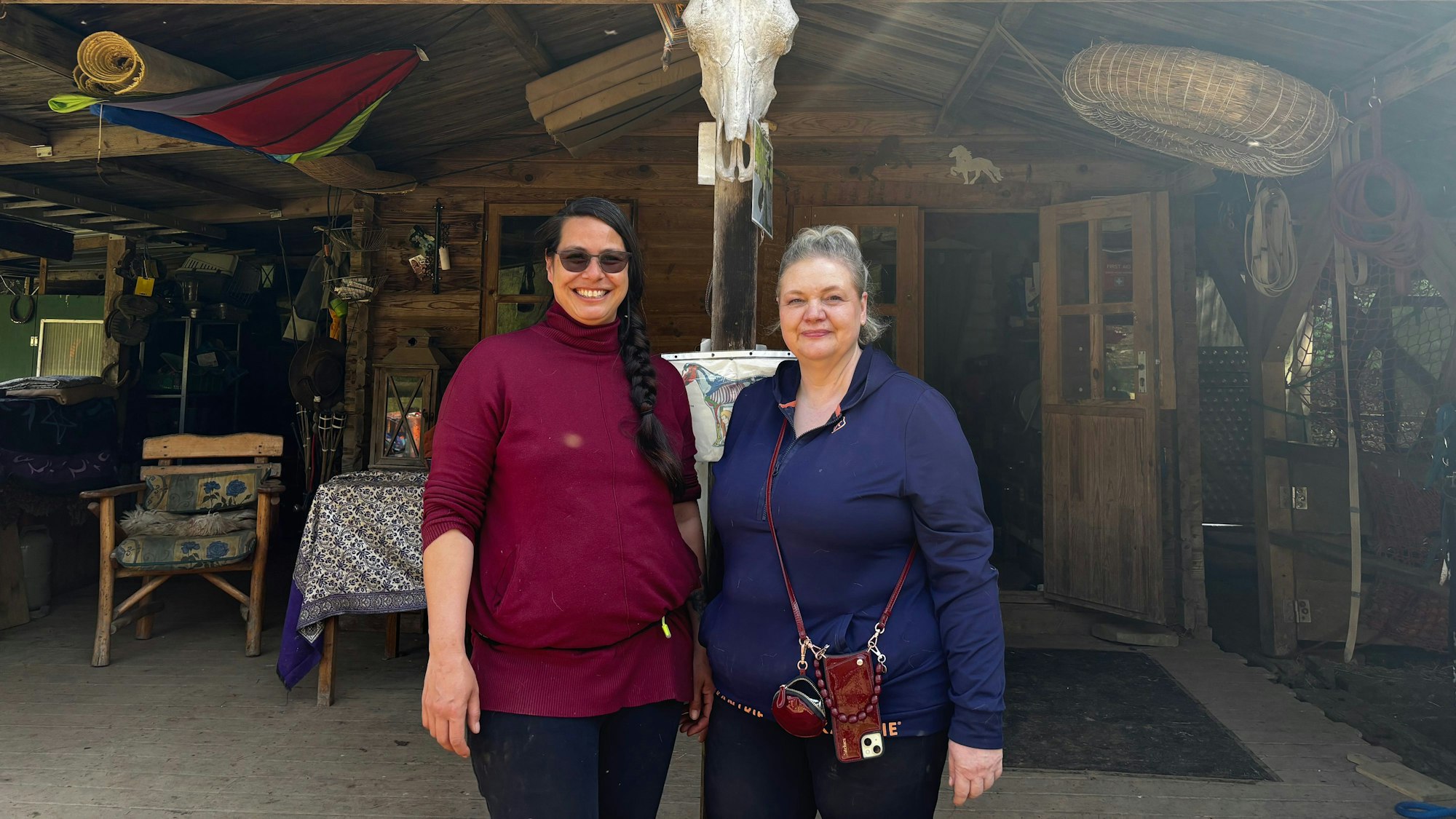 Zwei Frauen stehen nebeneinander und blicken lächelnd in die Kamera. Im Hintergrund ist die Ranch zu sehen.