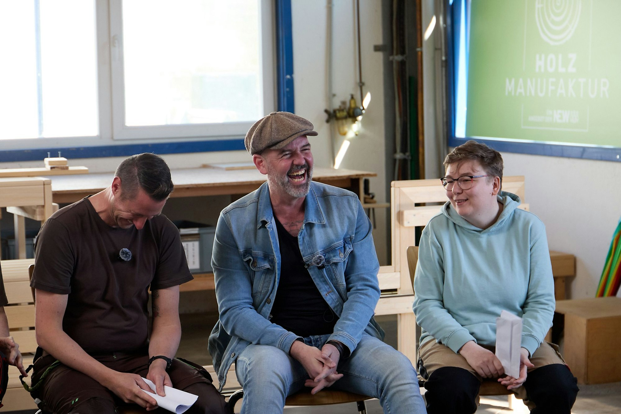 Doc Esser sitzt zwischen einem Mann und einer Frau in Arbeitskleidung und lacht herzlich.