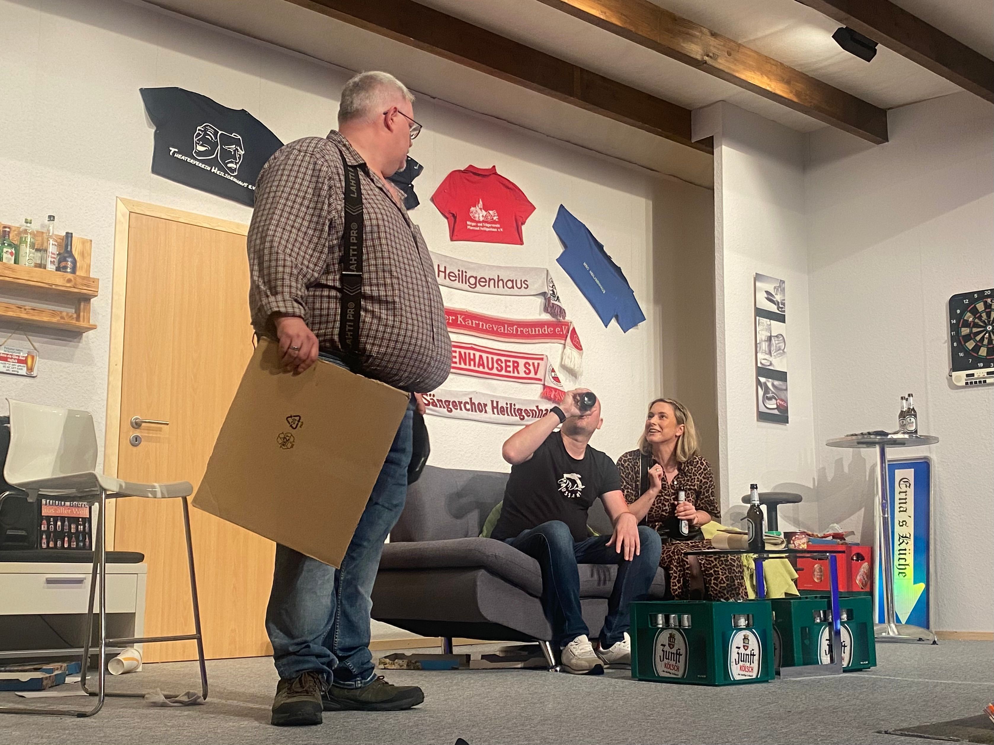 Auf einem Sofa sitzen zwei Personen, daneben steht ein Mann im Karohemd.