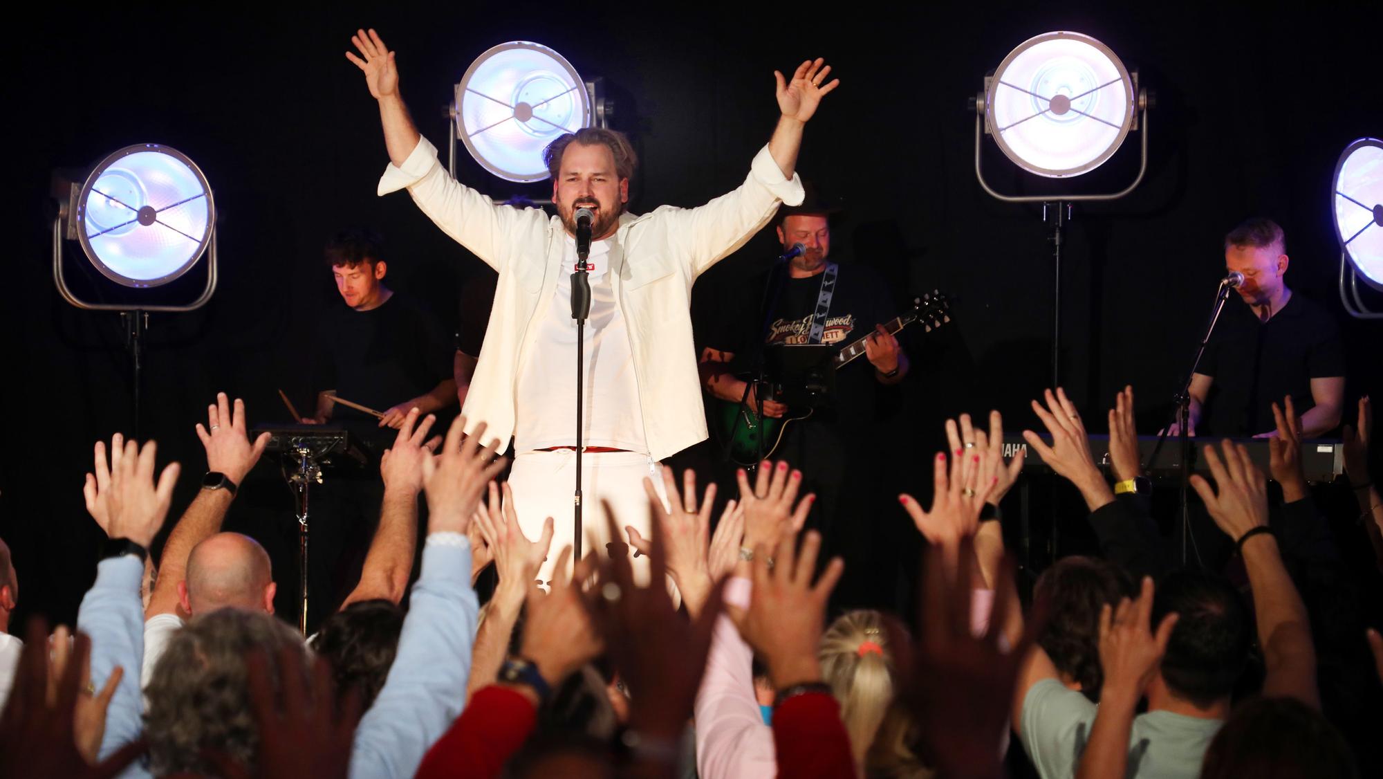 Ein Mann auf einer Bühne reckt die Hände in die Höhe so wie das Publikum vor ihm.