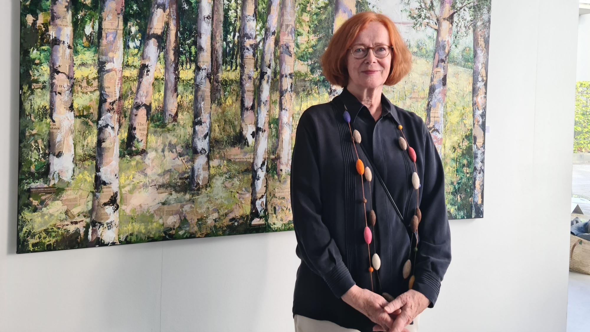 Eine Frau mit rotem Haar und Brille steht vor dem Bild eines Birkenwalds.