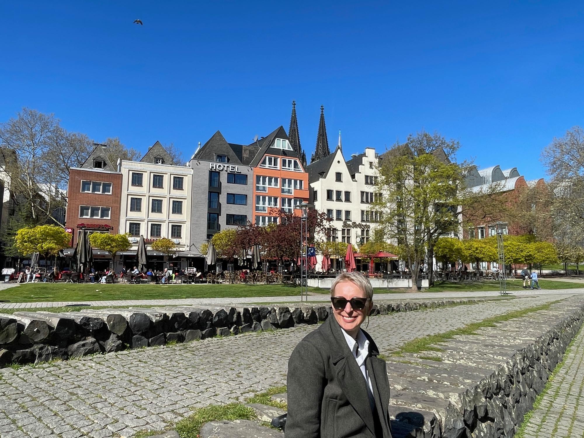 Eine Frau mit kurzem blonden Haar und Sonnenbrille sitzt auf einer Steinmauer. Hinter ihr sind die bunten Häuser der Kölner Altstadt zu sehen.