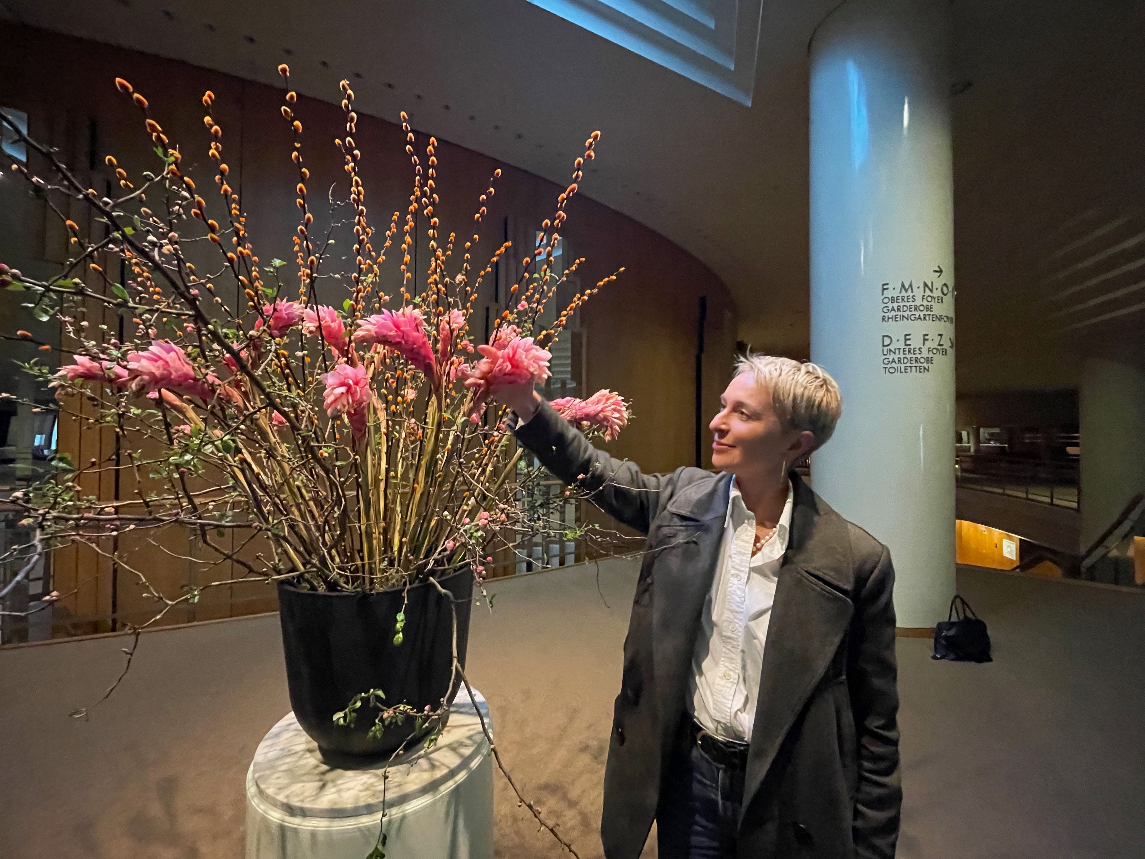 Eine Frau mit blondem, kurzem Haar im Anzug arrangiert Blumen in einer großen Vase in einem Foyer.