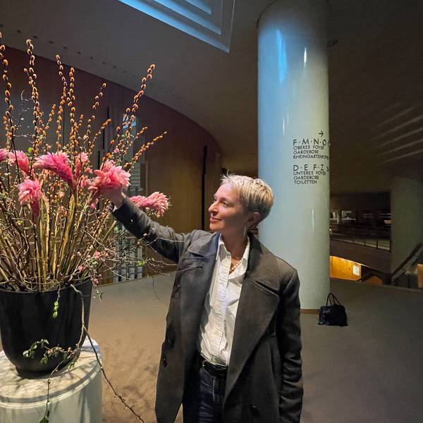 Eine Frau mit blondem, kurzem Haar im Anzug arrangiert Blumen in einer großen Vase in einem Foyer.