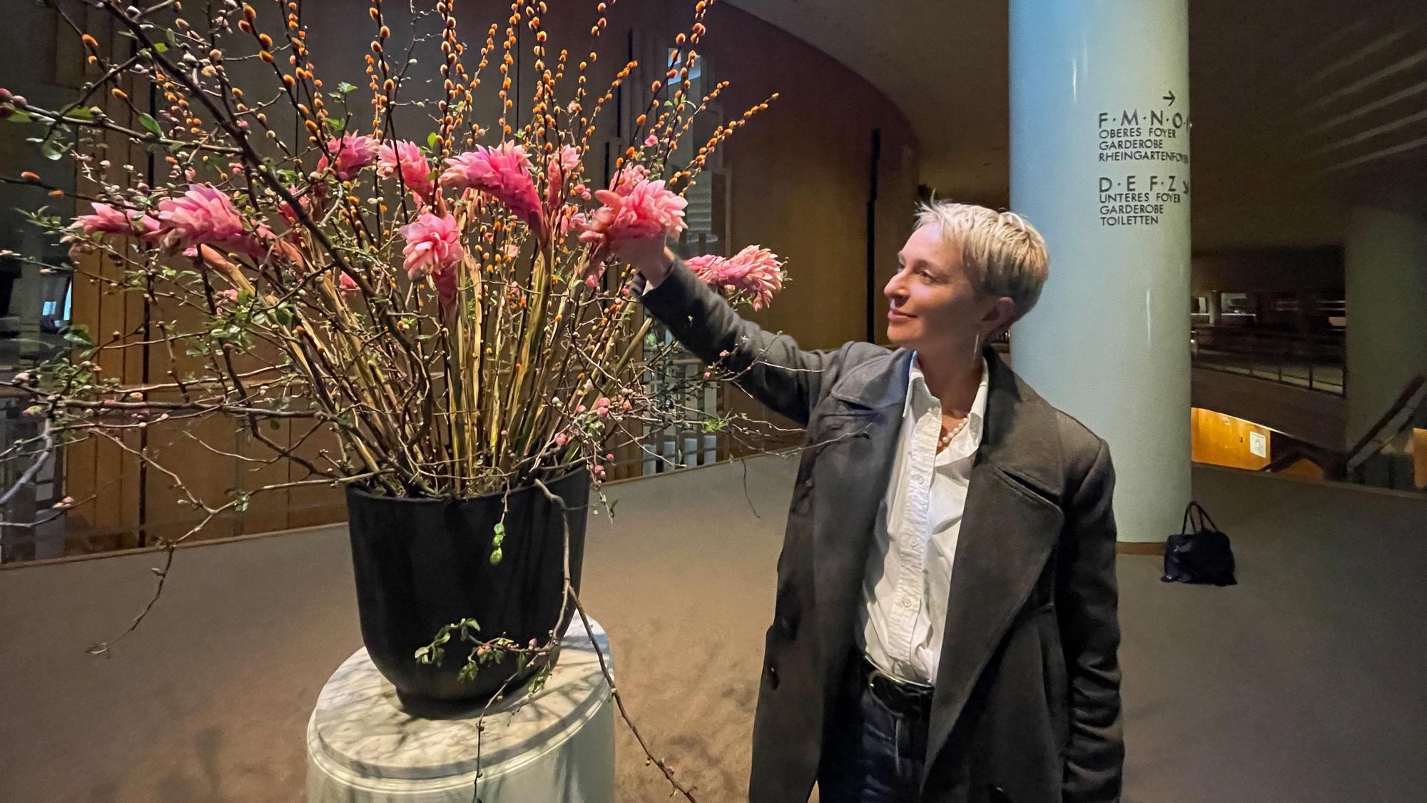 Eine Frau mit blondem, kurzem Haar im Anzug arrangiert Blumen in einer großen Vase in einem Foyer.