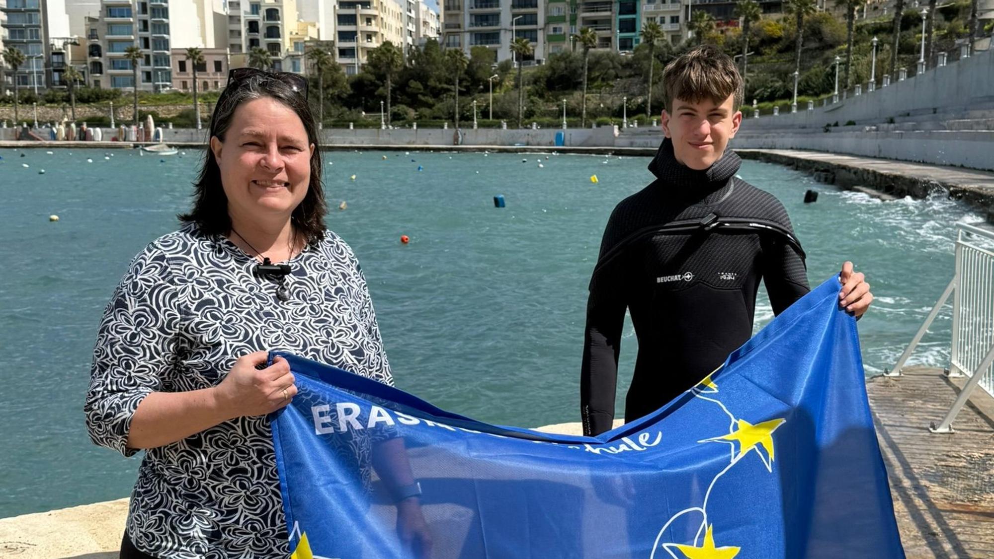 Eine Dame mittleren Alters und ein Schüler stehen am Meer und halten eine blaue Fahne in der Hand.