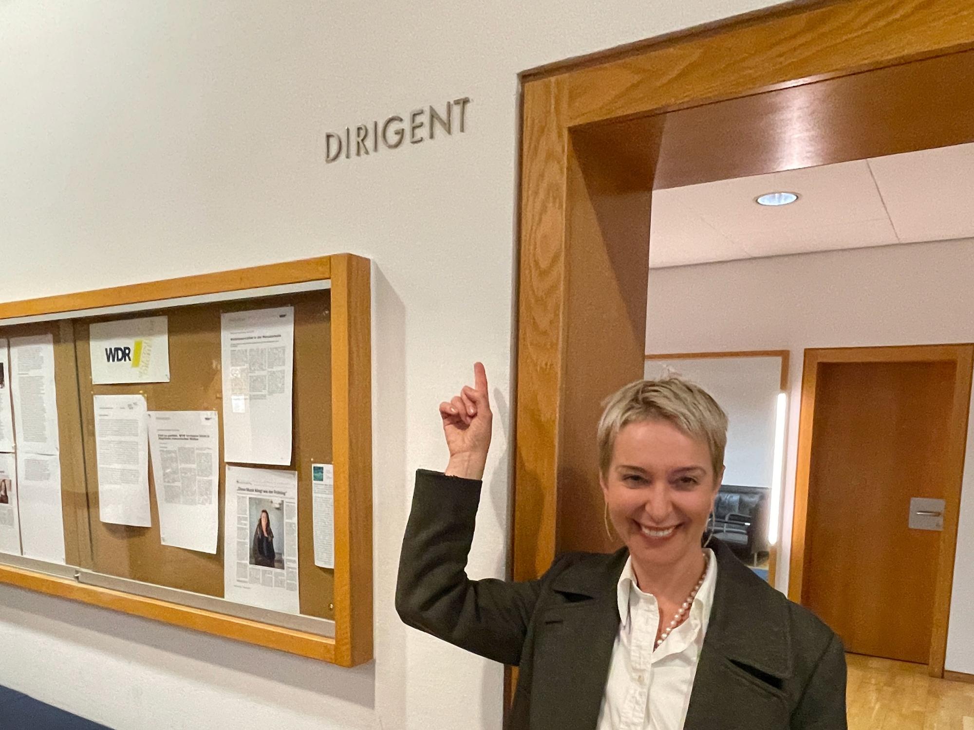 Eine Frau mit kurzem blonden Haar steht in einem Türrahmen. Sie zeigt auf den Schriftzug „Dirigent“, der oben neben der Tür prangt.