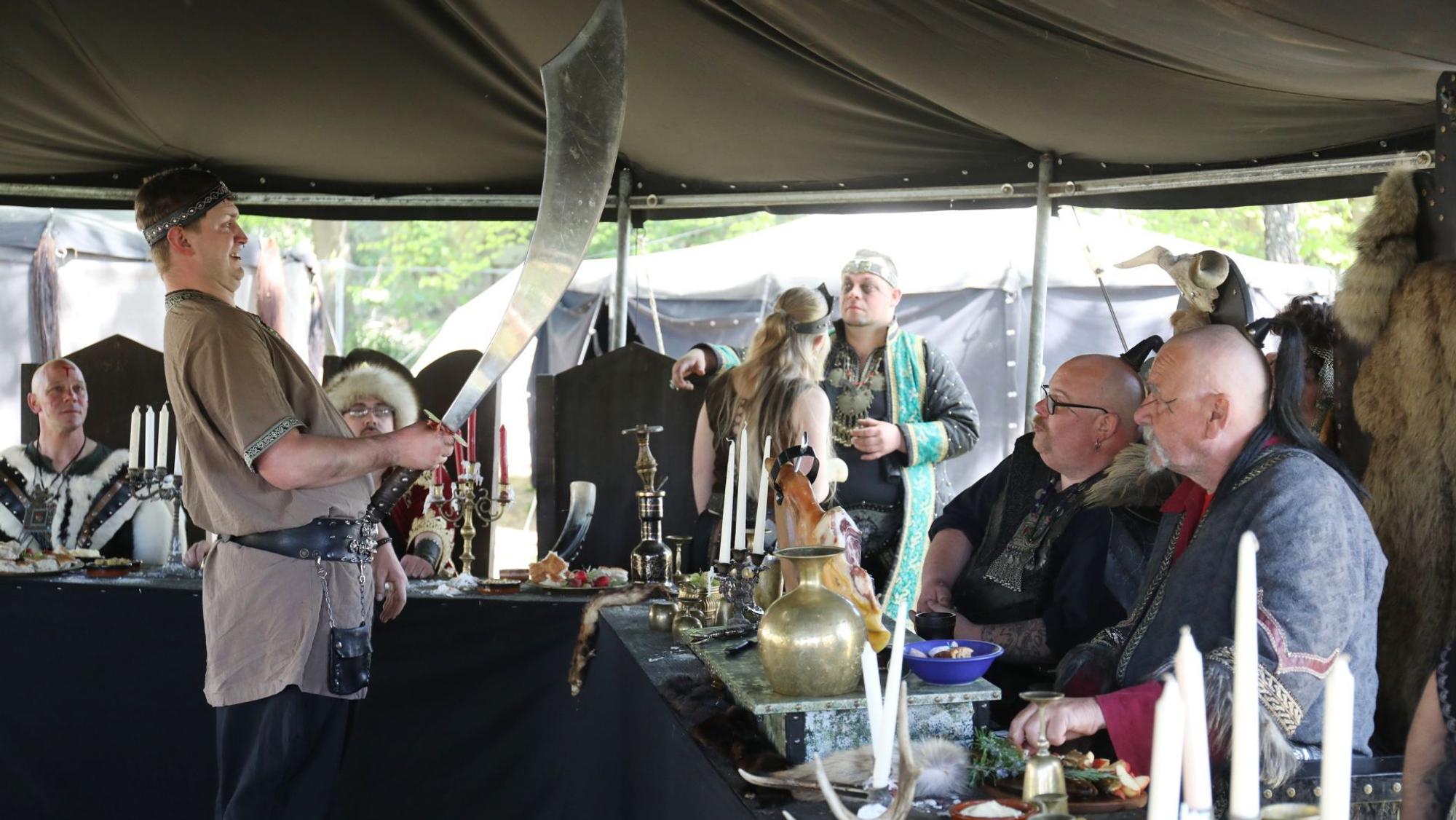 Eine Gruppe von Menschen, die als Hunnen verkleidet sind, an einer Tafel. Einer hebt ein schweres Schwert.