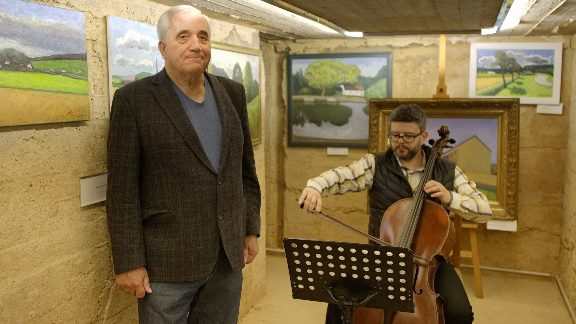 Ein älterer Mann steht vor Bildern, während neben ihm ein jüngerer Mann auf einem Bass musiziert.