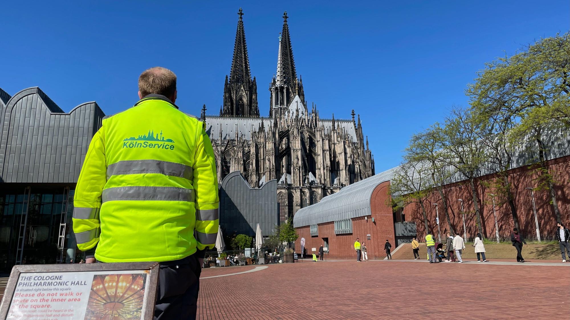 Ein Mann in gelber Warnweste steht auf einem Platz, im Hintergrund sind das Museum Ludwig und der Dom zu sehen.