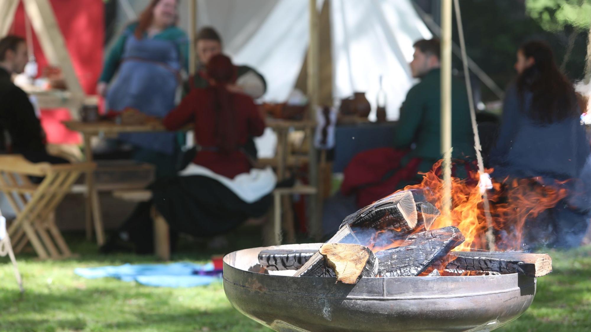 Eine Feuerschale mit brennenden Holzscheiten, im Hintergrund Menschen vor Zelten.