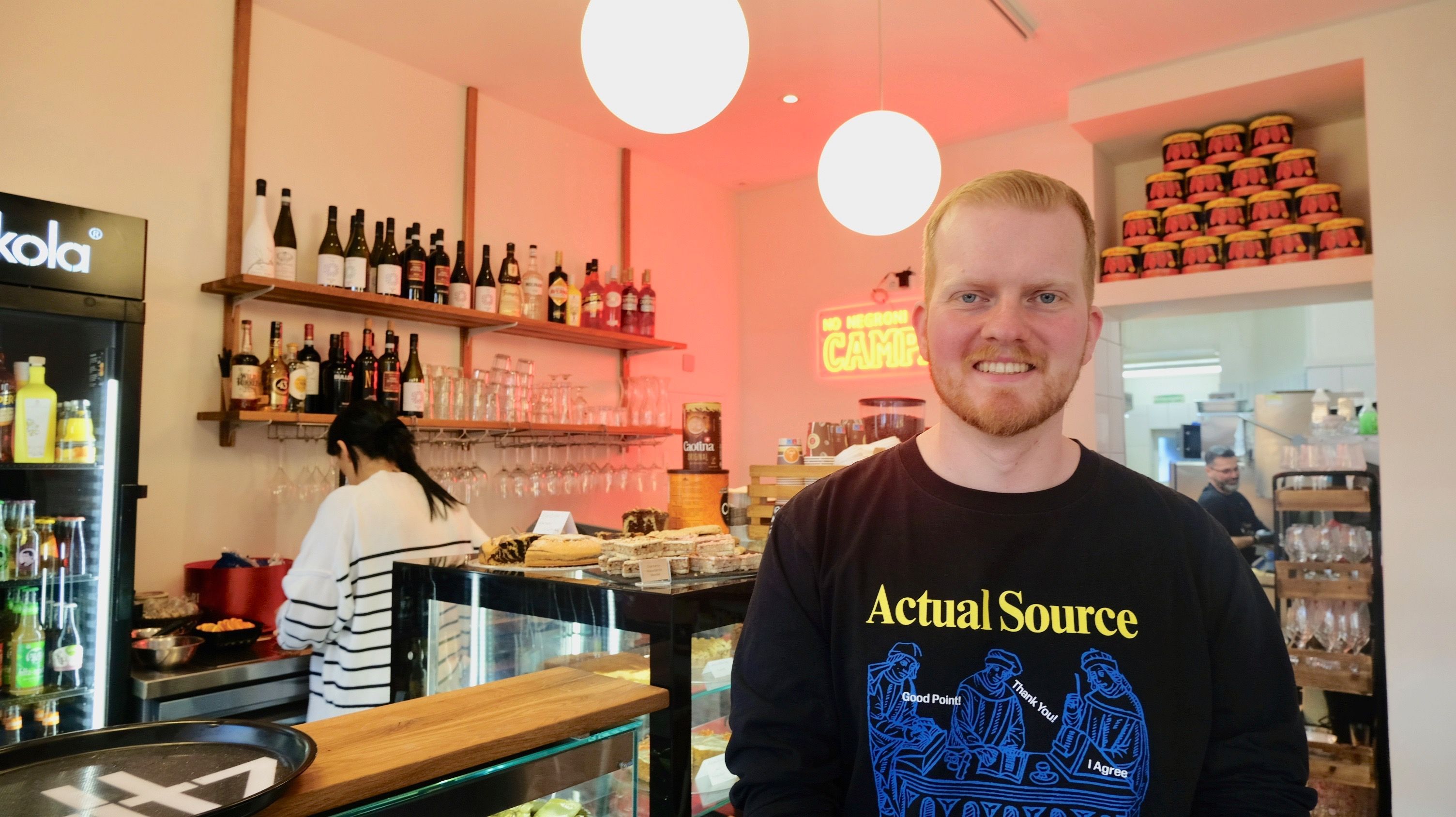 Ein junger Mann mit kurzem, rotblonden Haar und Bart steht vor einer Kuchentheke in einem Café.