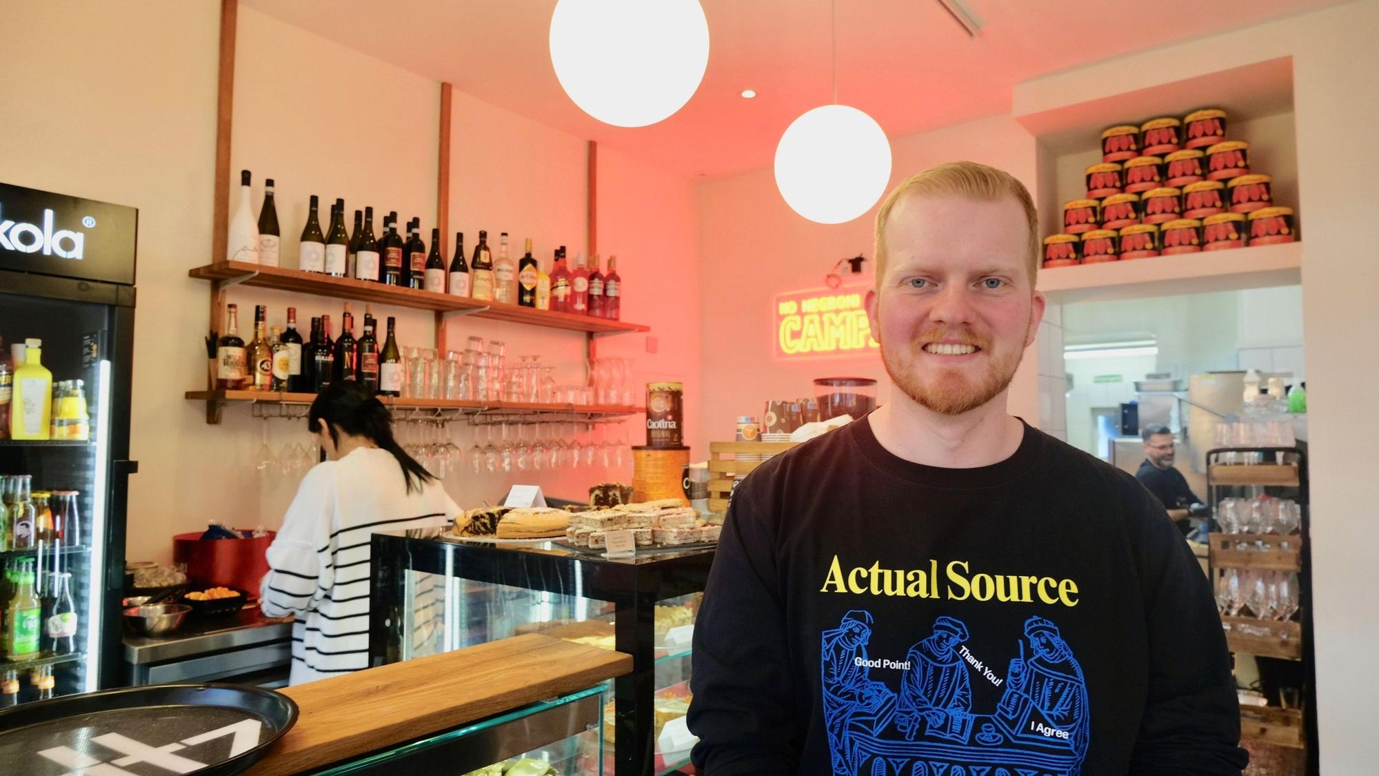 Ein junger Mann mit kurzem, rotblonden Haar und Bart steht vor einer Kuchentheke in einem Café.