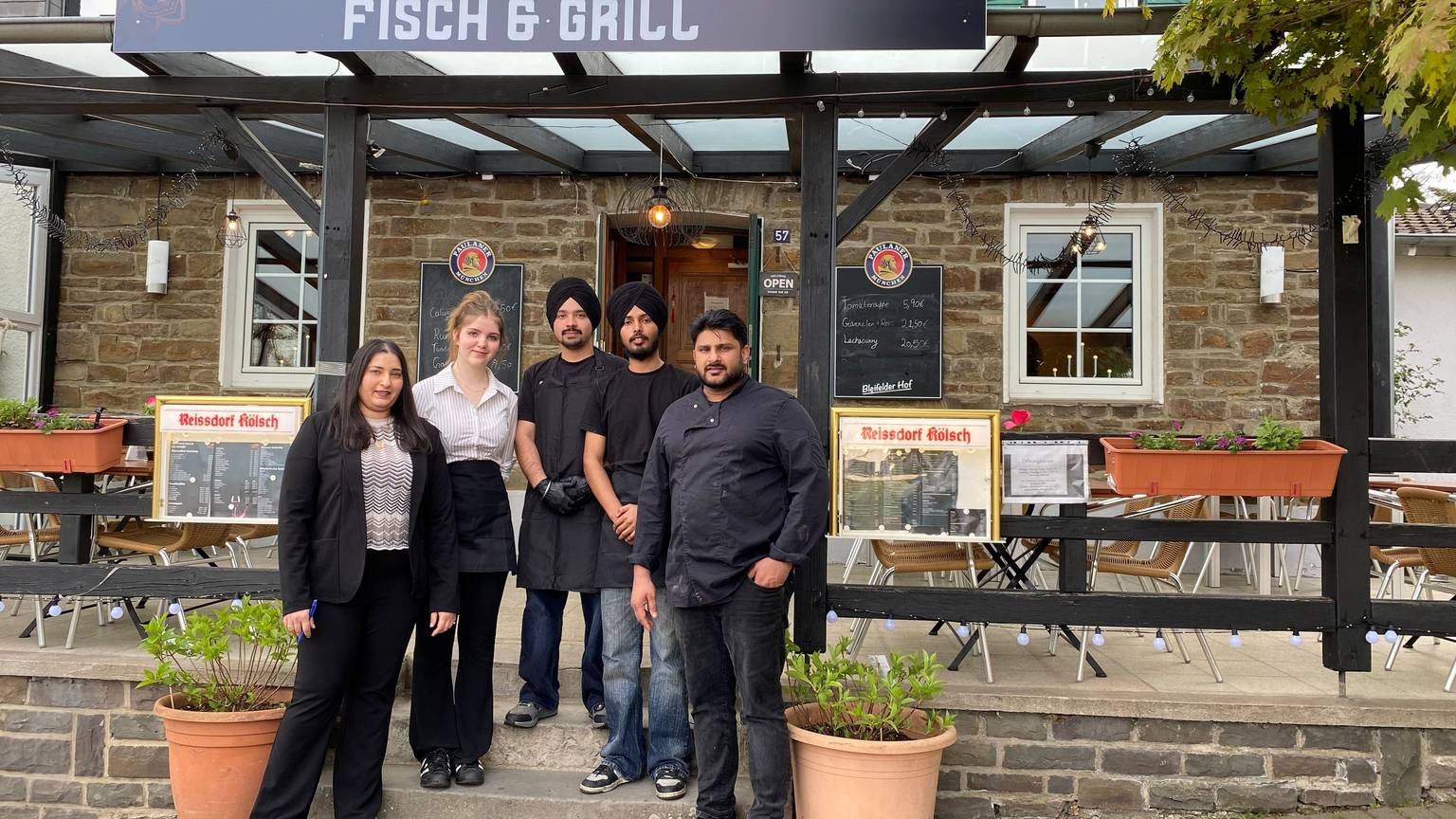Oben das Schild des Restaurants mit seinem Namen, davor die beiden Inhaber, eine junge Frau aus Bleifeld und zwei junge Männer aus Indien, beide mit schwarzem Turban.
