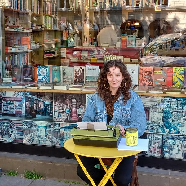 Eine junge Frau mit lockigem Haar sitzt an einem kleinen gelben Tisch vor einer Buchhandlung. Sie schreibt auf einer grünen Schreibmaschine.