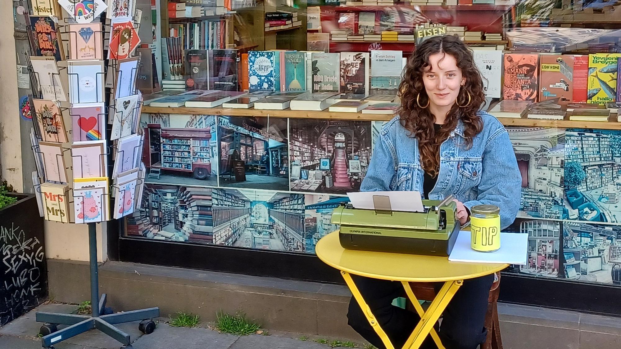 Eine junge Frau mit lockigem Haar sitzt an einem kleinen gelben Tisch vor einer Buchhandlung. Sie schreibt auf einer grünen Schreibmaschine.