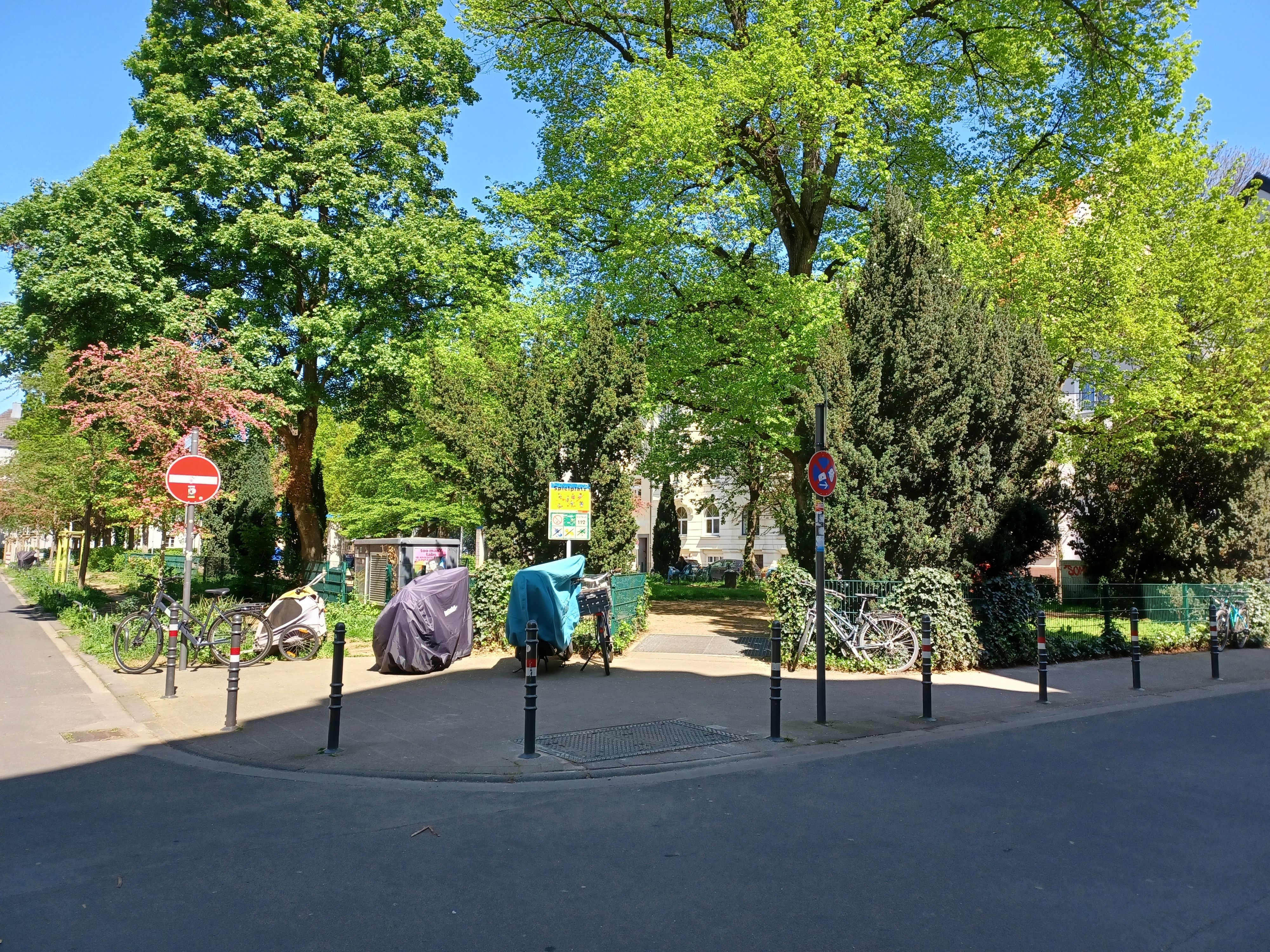 Blick auf einen Platz mit hohen Bäumen, am Rand stehen Poller, dahinter führt ein Weg auf die Platzfläche.