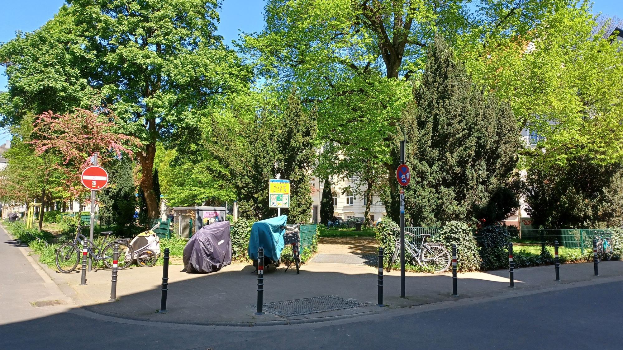 Blick auf einen Platz mit hohen Bäumen, am Rand stehen Poller, dahinter führt ein Weg auf die Platzfläche.