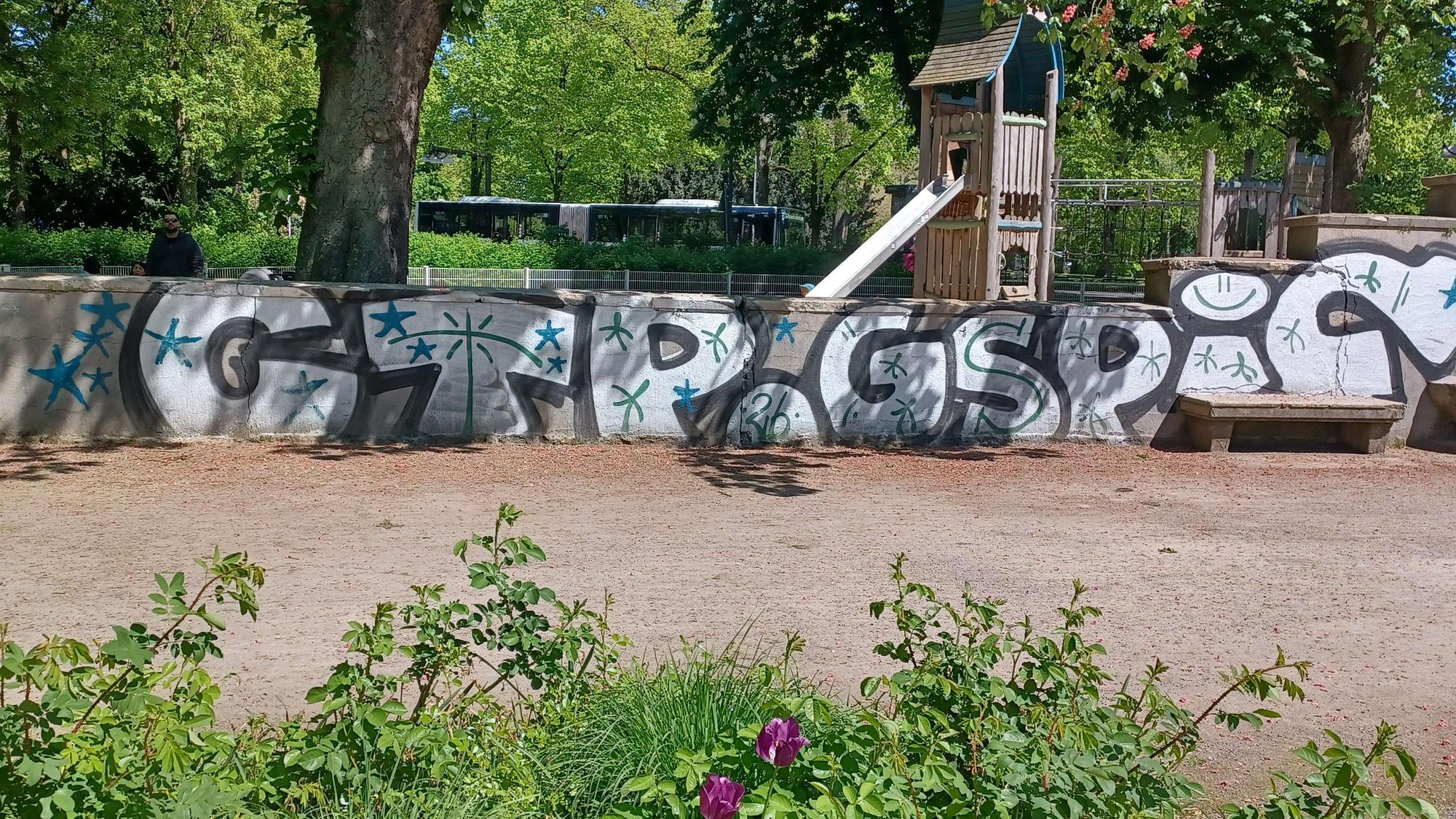Blick auf eine mit Graffiti besprühte Mauer, dahinter liegt ein Spielplatz.