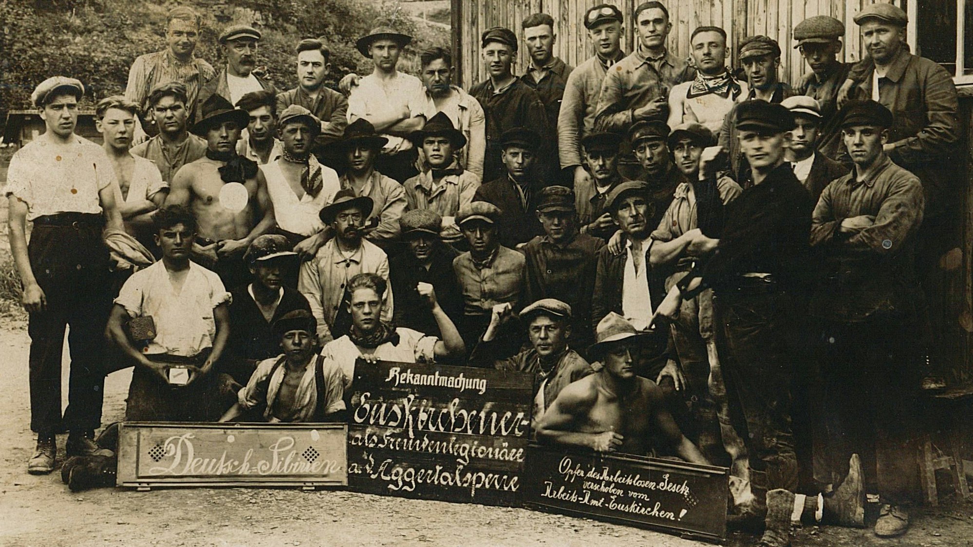 Dies ist die Vorderseite einer nicht gelaufenen Postkarte. Die Fotografie zeigt offensichtlich unzufriedene Arbeiter aus dem Raum Euskirchen, die sich laut einem Schild als „Opfer des Arbeitslosengesetzes“ sehen, beim Bau der Aggertalsperre bei Gummersbach, vermutlich 1927.