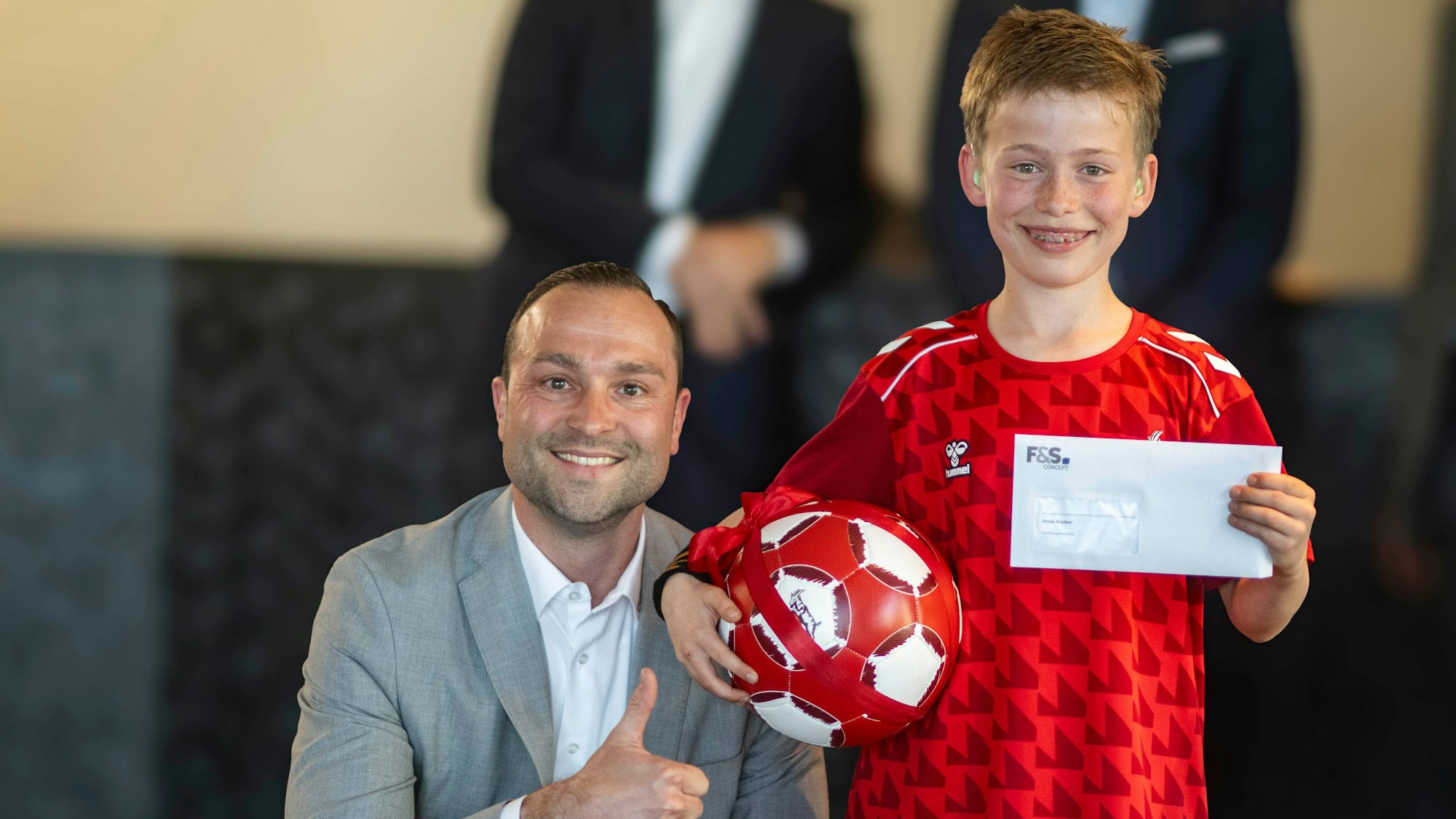 Jan Radermacher kniet neben Jonas Klaiber, der einen Fußball unterm Arm und einen Umschlag in der Hand hält.
