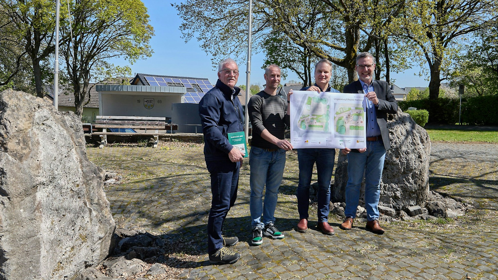 Vier Männer stehen auf dem Eiffelplatz in Nettersheim und halten gemeinsam eine große Planungsskizze.