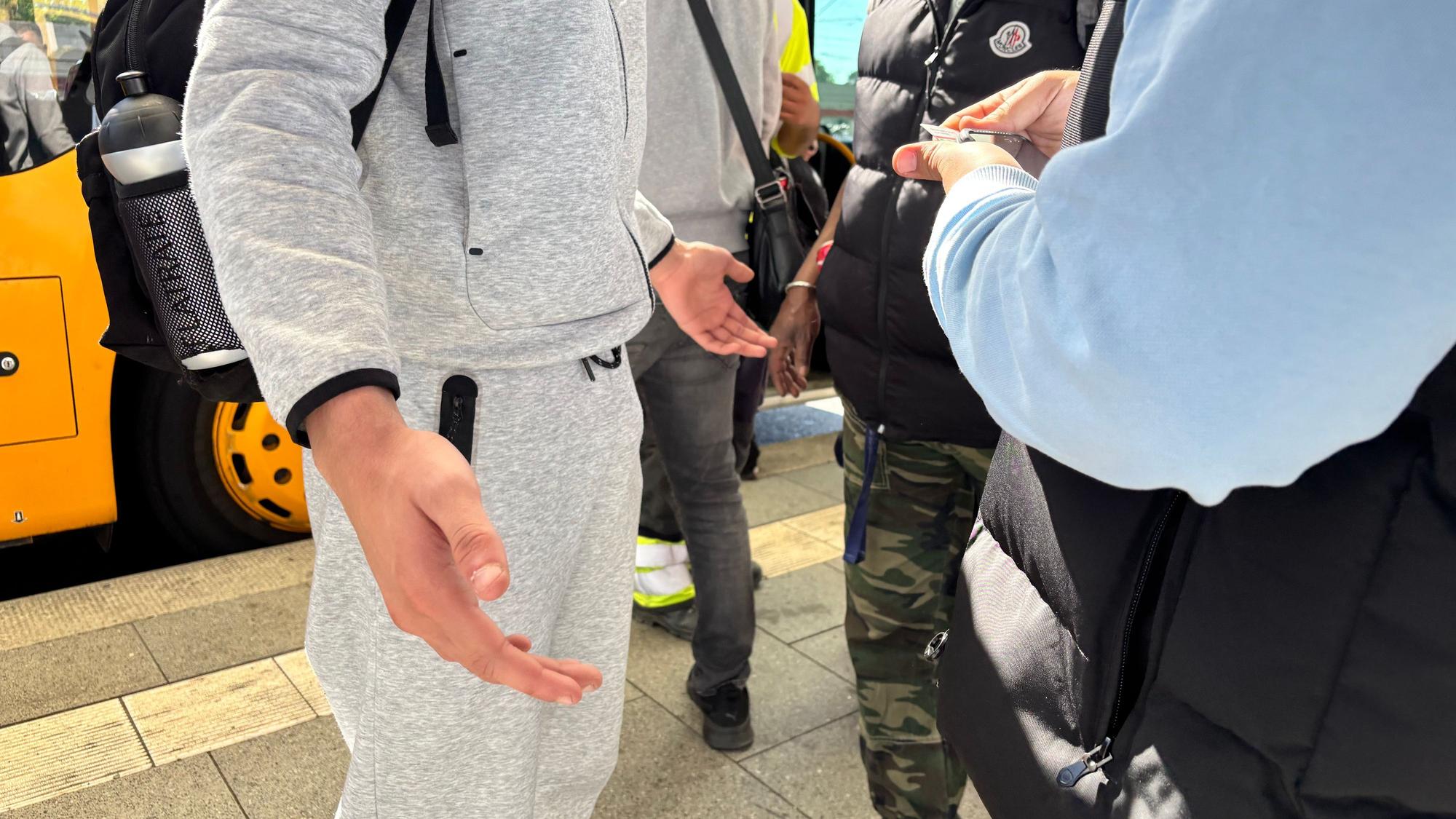 Eine Person in einem grauen Trainingsanzug macht eine ausladende Geste mit beiden Händen, während ein Kontrolleuer vor ihr steht. Die Gesichter der Beteiligten sind abgeschnitten.