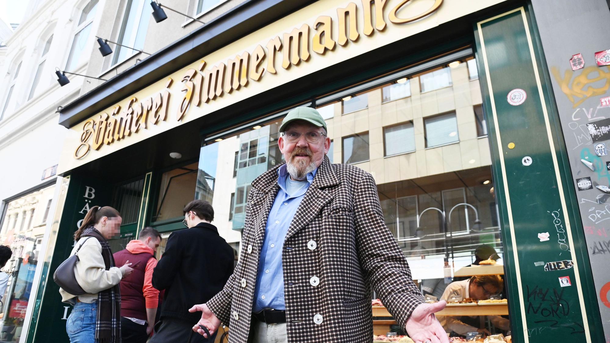 Ein Mann im karierten Sakko mit grüner Kappe und Bart steht mit ausgebreiteten Armen vor einer Bäckerei.