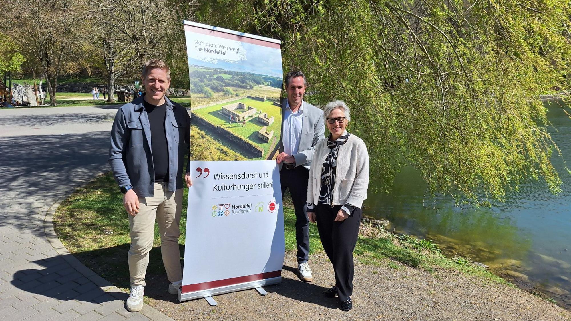 Die drei Genannten vor einem Plakat, das für den Tourismus in der Nordeifel wirbt. Im Hintergrund ist der Blankenheimer Weiher zu sehen.
