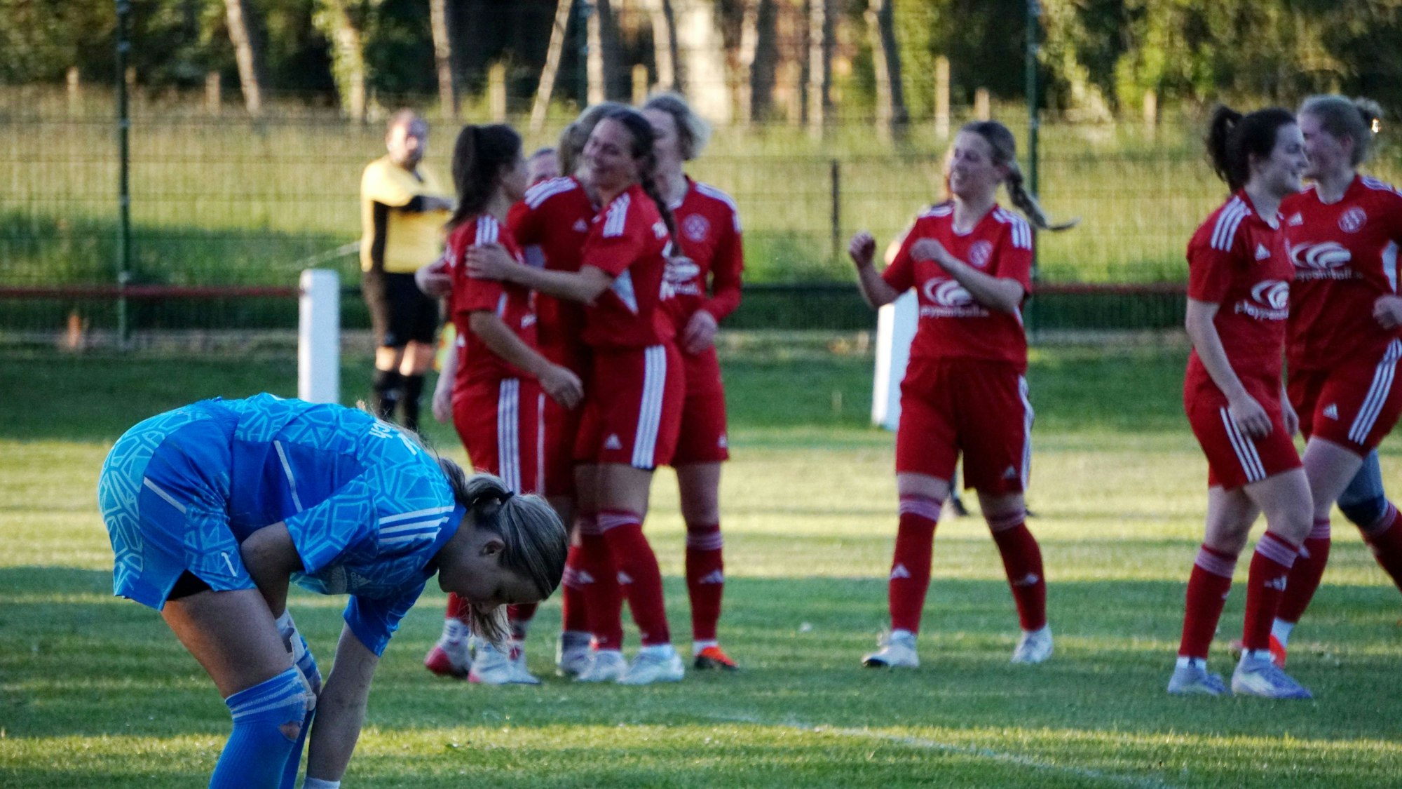 Im Hintergrund jubelt eine Fußballmannschaft, im Vordergrund beugt sich die gegnerische Torhüterin nach vorne