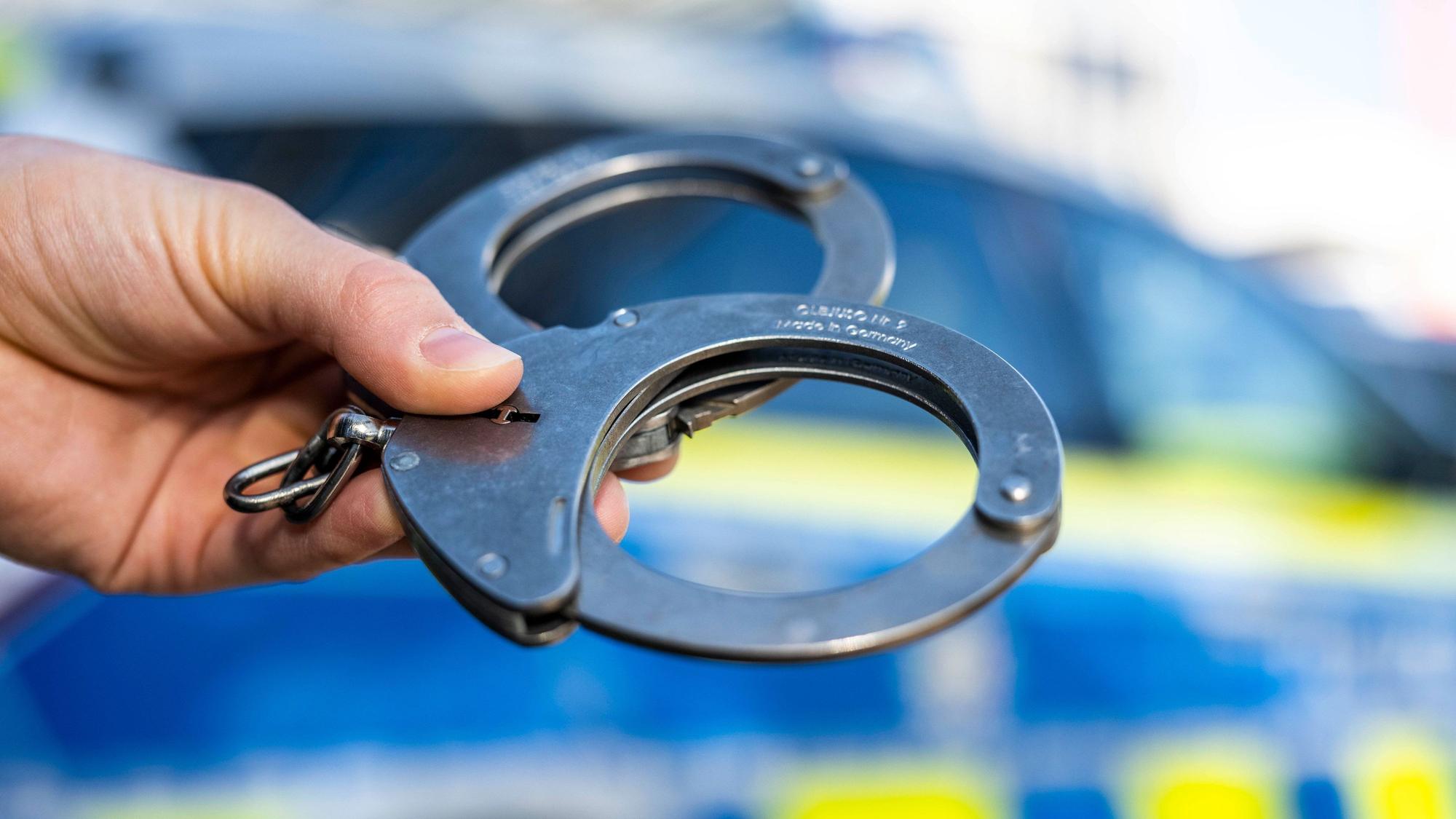 Eine Hand hält Handschellen vor einen Streifenwagen der Polizei. (Symbolbild)