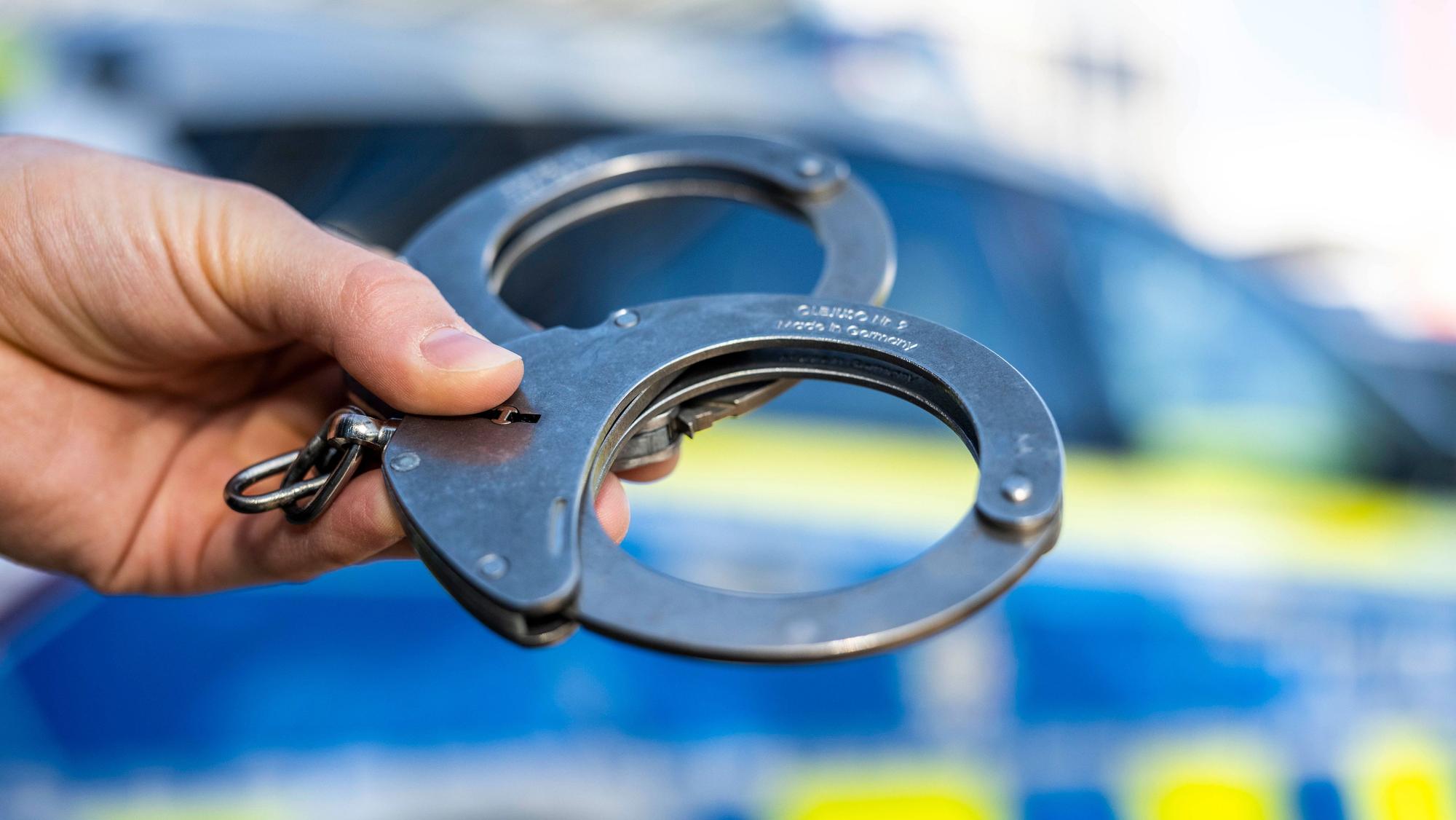 Eine Hand hält Handschellen vor einen Streifenwagen der Polizei. (Symbolbild)