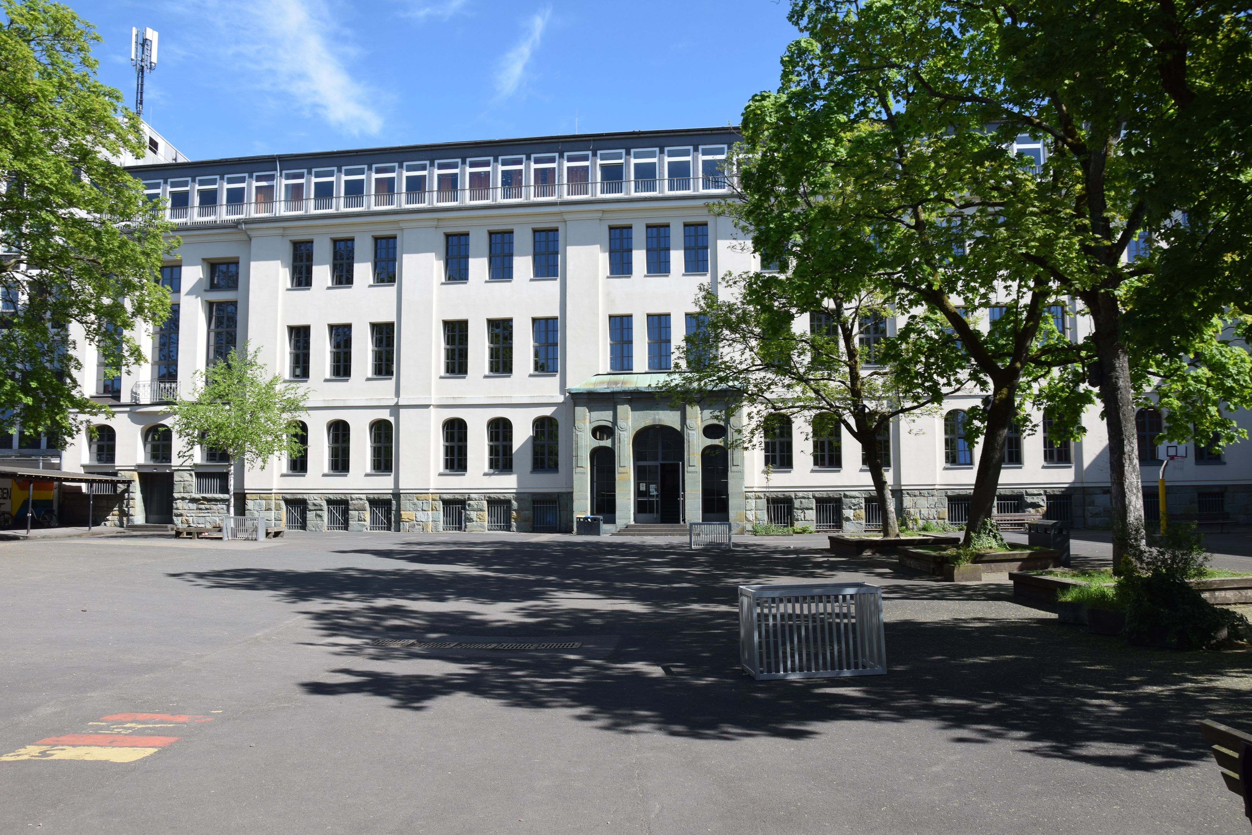 Hinter einem Schulhof steht ein mehrtöckiges weiß getünchtes Gebäude.