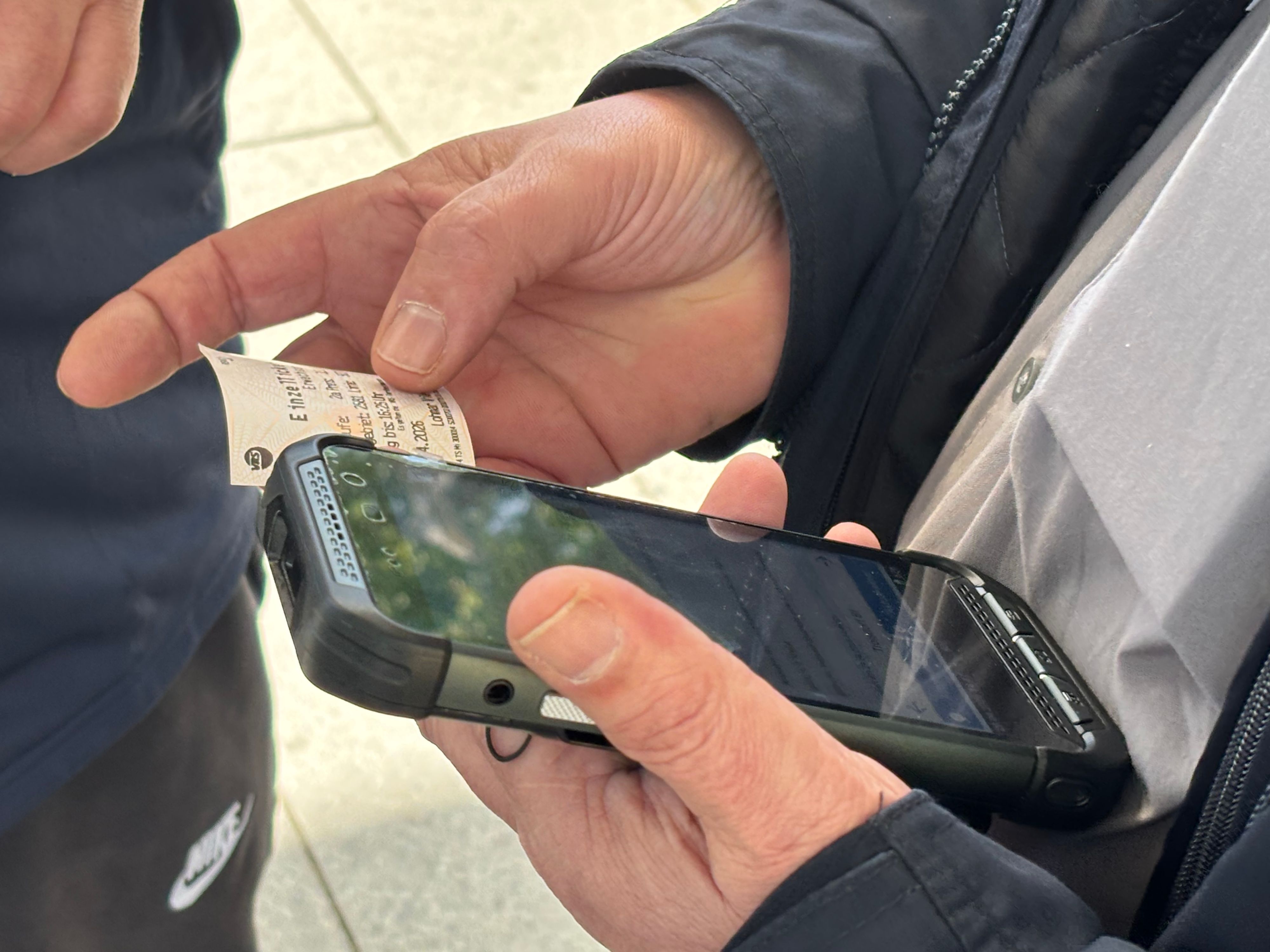 Die Nahaufnahme zeigt Zwei Hände: In der linken Hand ruht ein Handy, die rechte Hand hält eine Papier-Fahrkarte daneben.