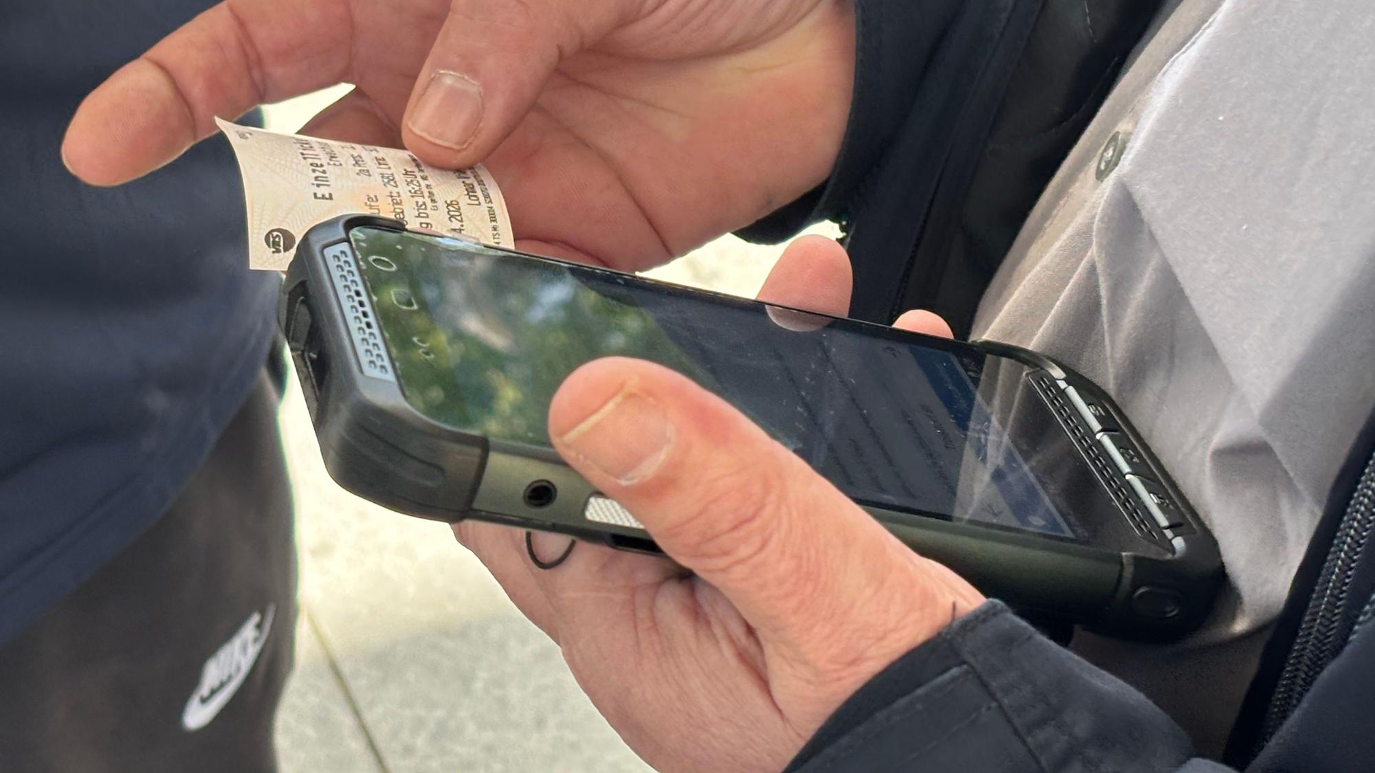 Die Nahaufnahme zeigt Zwei Hände: In der linken Hand ruht ein Handy, die rechte Hand hält eine Papier-Fahrkarte daneben.