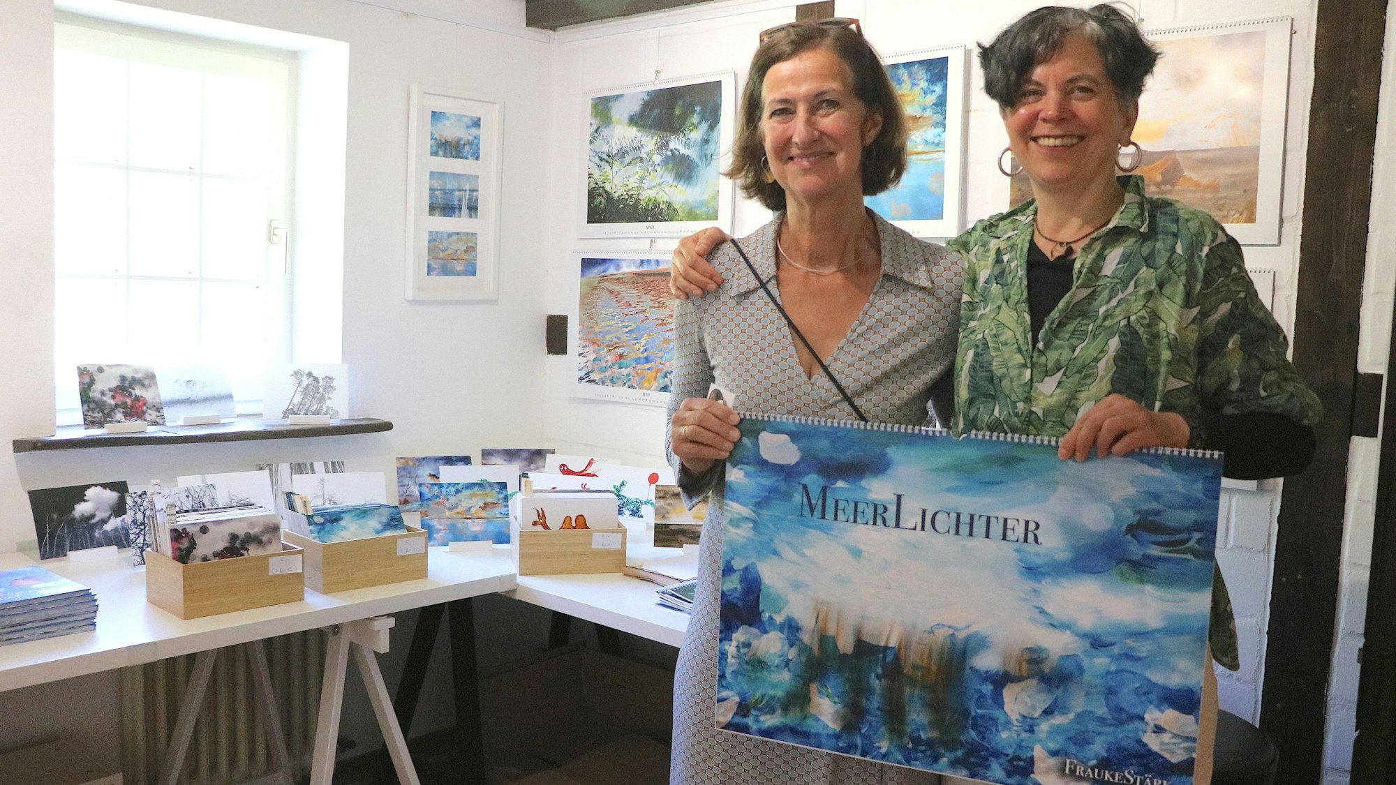 Zwei Frauen stehen nebeneinander, sie halten einen blauen Wandkalender in den Händen.