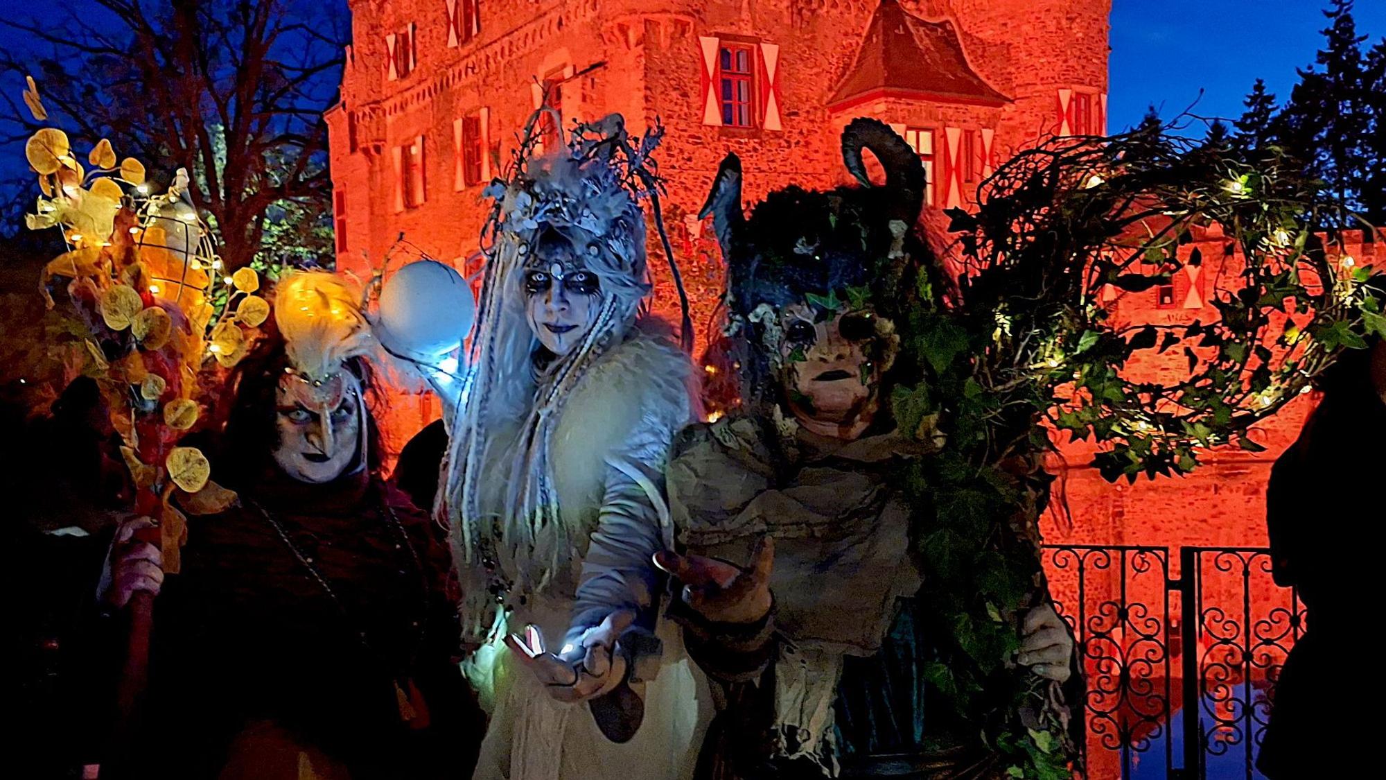 Die drei kostümierten Hexen in aufwendigen Kostümen vor der orange angestrahlten Burg.
