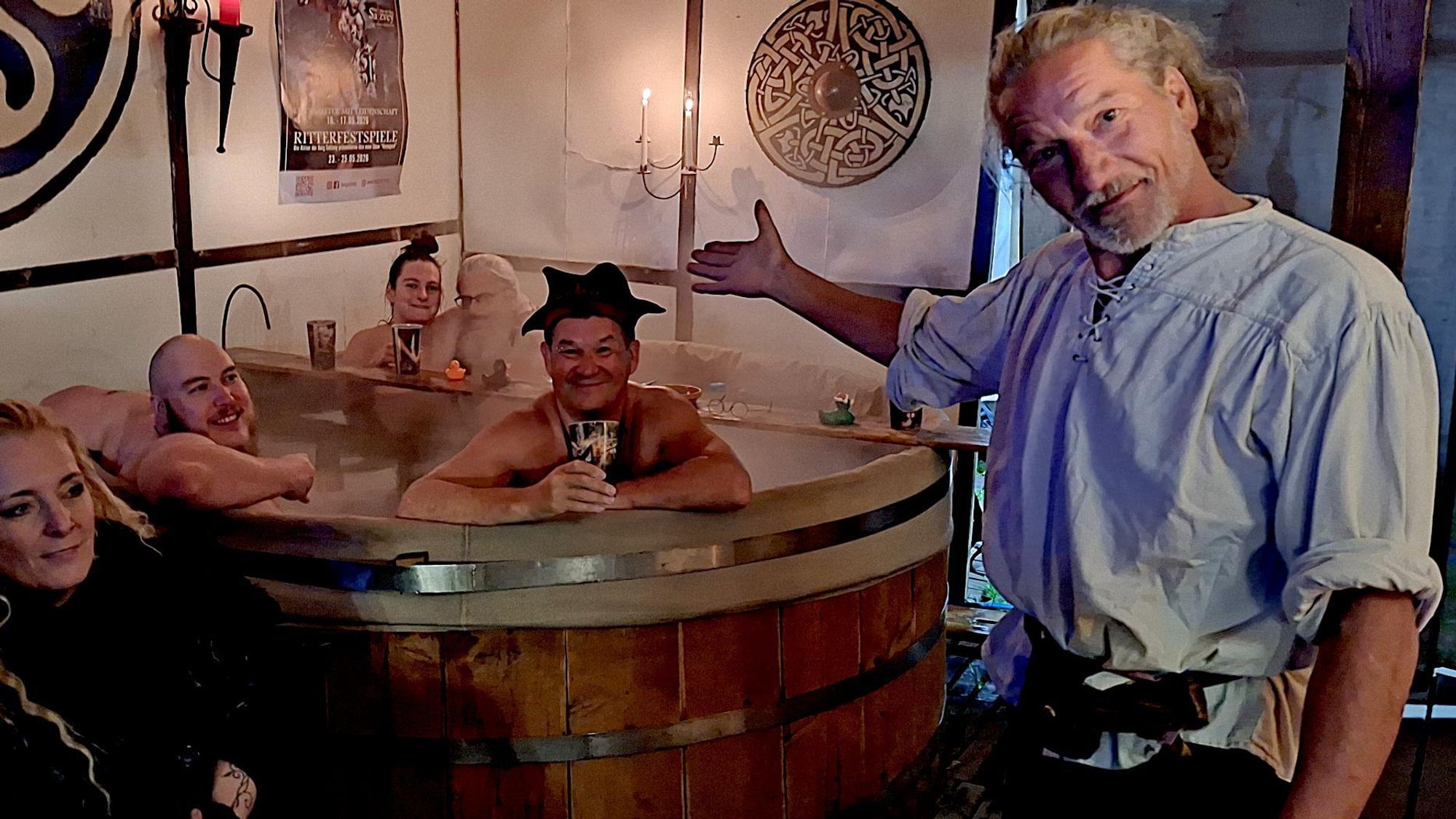 Michael Jansen weist auf den hölzernen Badezuber, in dem vier Badegäste sitzen.