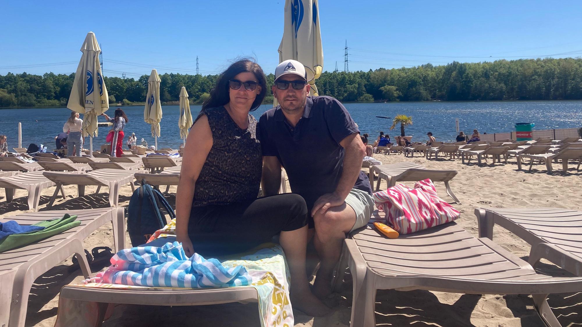 Eine Frau und ein Mann sitzen auf Strandliegen, im Hintergrund ein See.