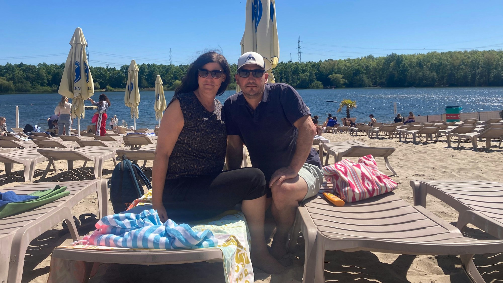 Eine Frau und ein Mann sitzen auf Strandliegen, im Hintergrund ein See.