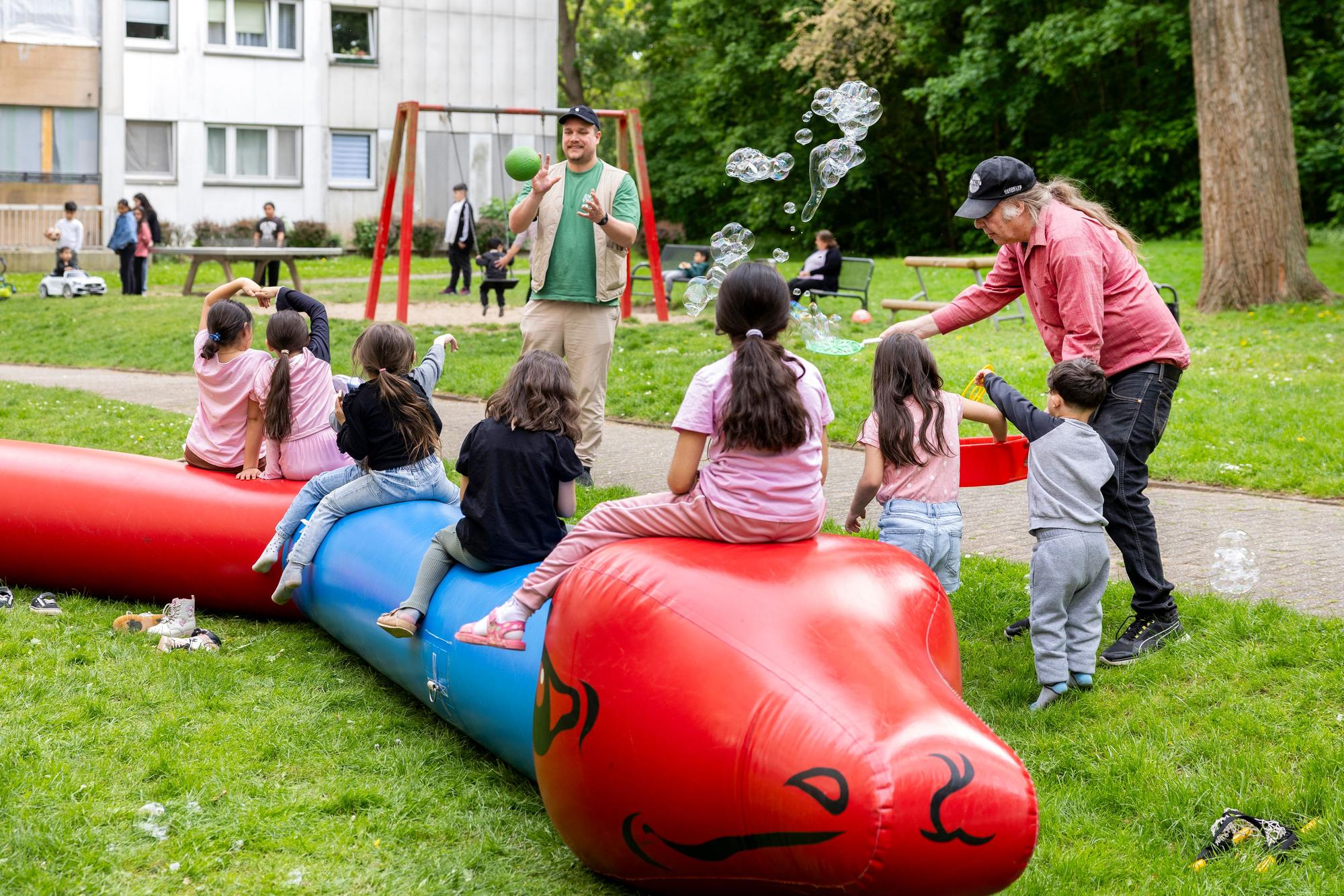 Kinder sitzen auf einer langen Hüpfschlange im Hinterhof der tristen Hochhaussiedlung in der Gernsheimer Straße. Mitarbeiter der Kölner Spielewerkstatt unterhalten die Kinder mit Seifenblasen und Ballspielen.