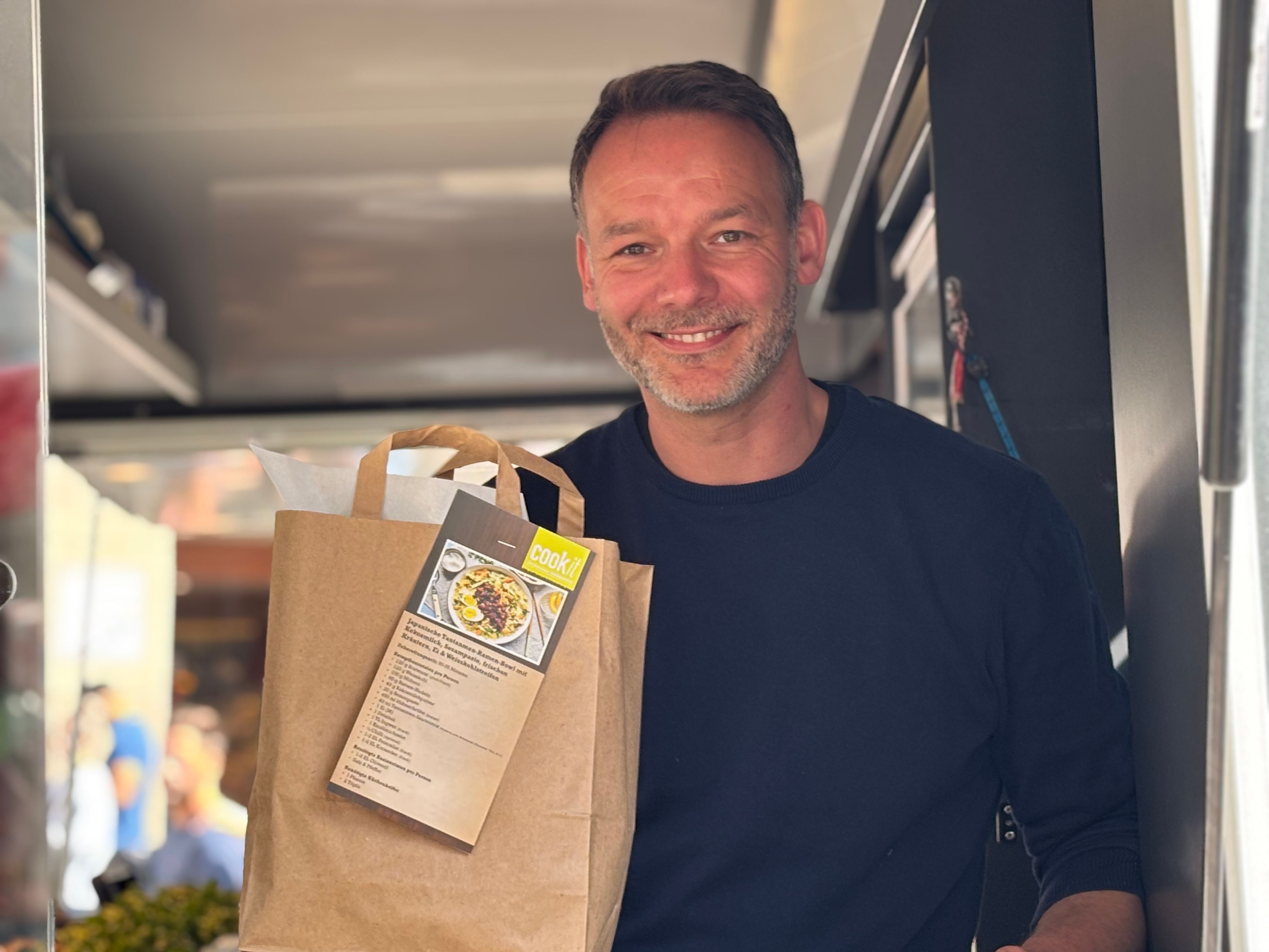 Tim Engelbrecht steht in einem fahrbaren Marktstand und hält lächelend eine Kochtüte in die Kamera.