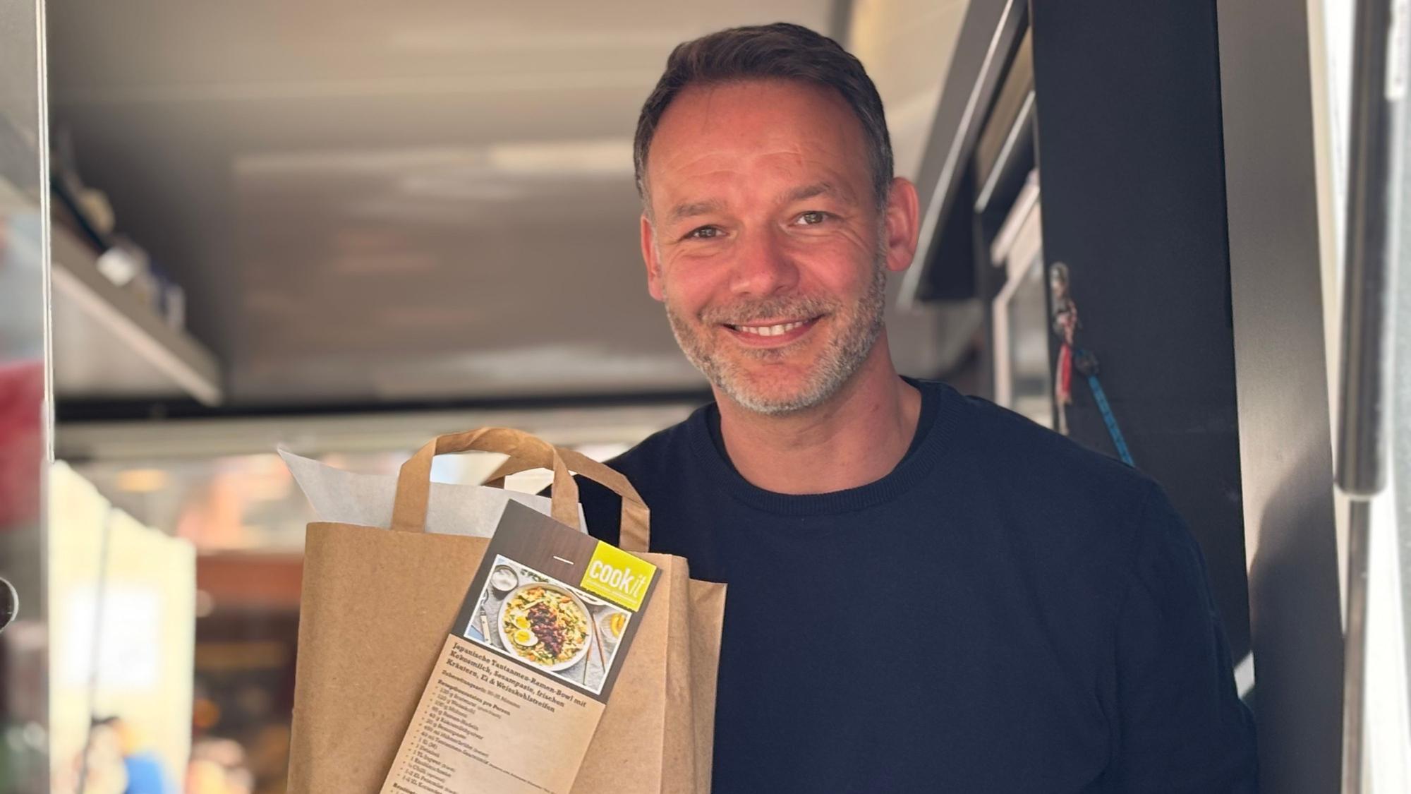 Tim Engelbrecht steht in einem fahrbaren Marktstand und hält lächelend eine Kochtüte in die Kamera.